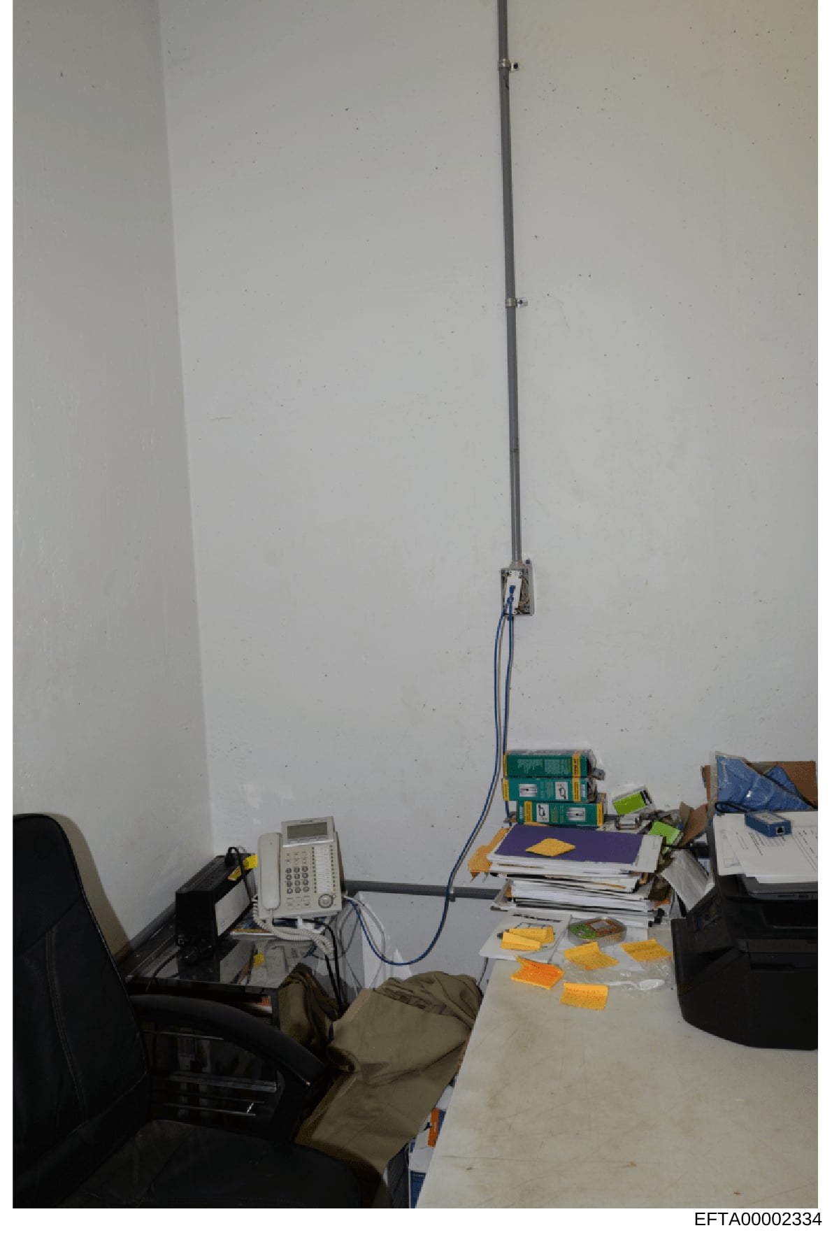 This is an evidence photograph showing an interior office or workspace containing various electronic equipment, documents, and materials. The image captures a desk area with a telephone/communications device, stacks of papers and files, storage boxes
