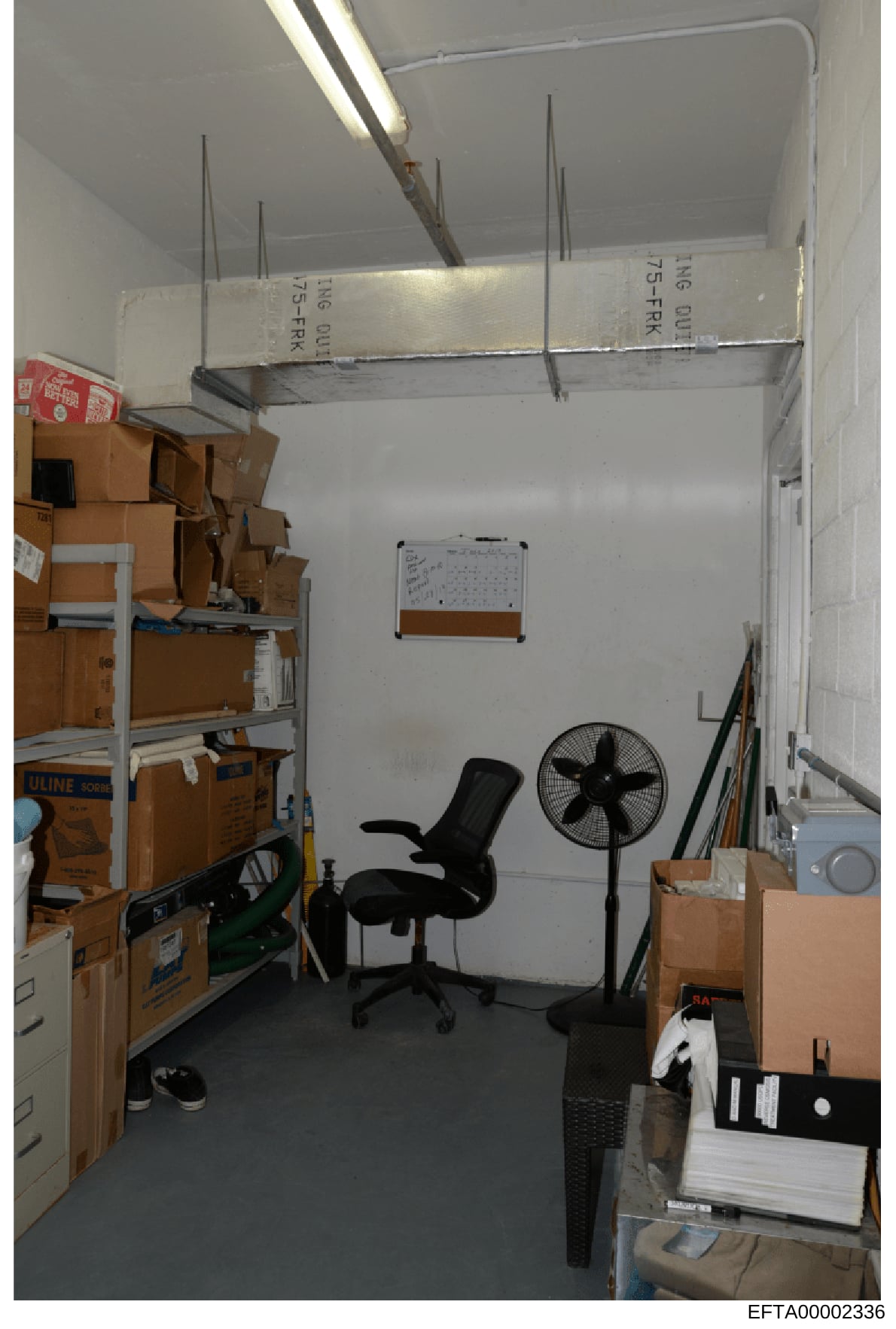 This is an evidence photograph showing the interior of a room, likely a storage area or office space, containing multiple cardboard boxes, office furniture, and equipment. The room appears to be documented as part of the investigation's evidence coll