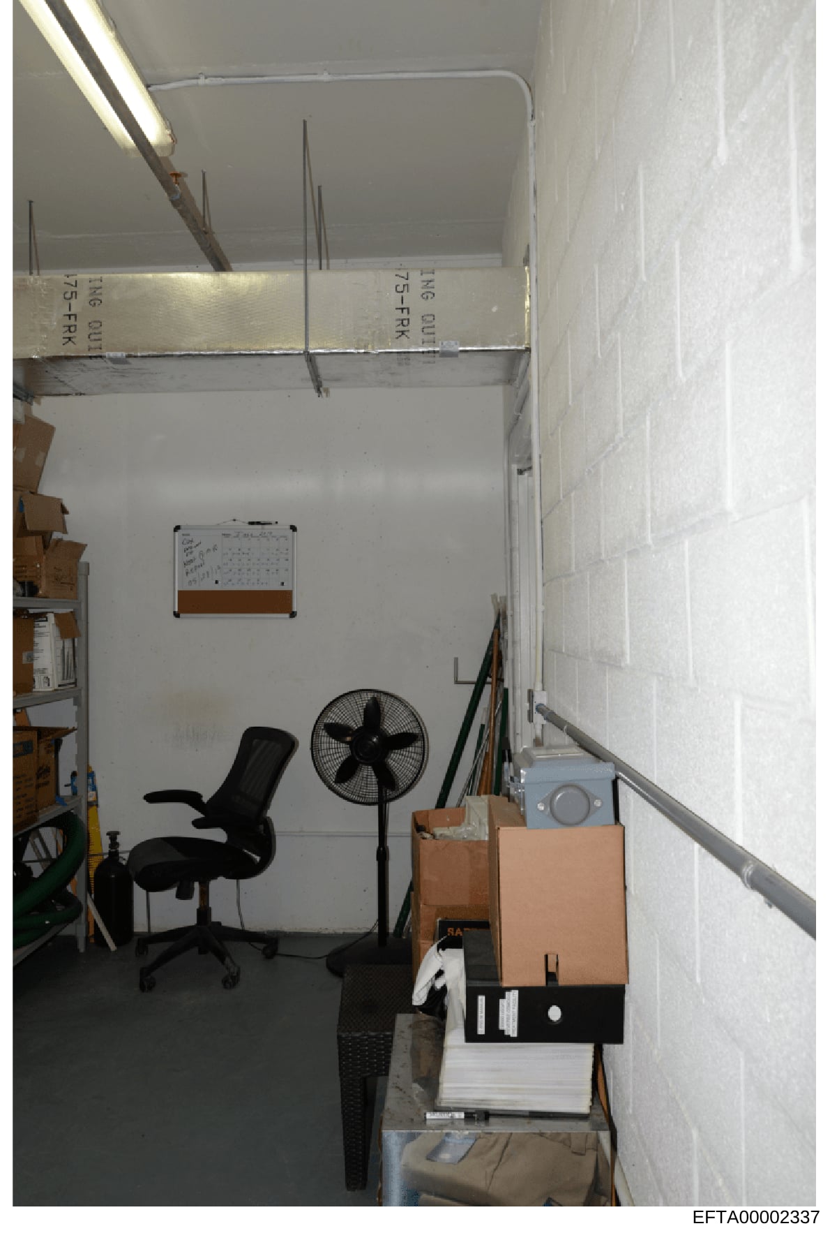 This is an evidence photograph showing an interior room with storage boxes, office furniture, and various items. The image appears to be taken during a property search or investigation, with labeled boxes visible at the top of the frame bearing marki