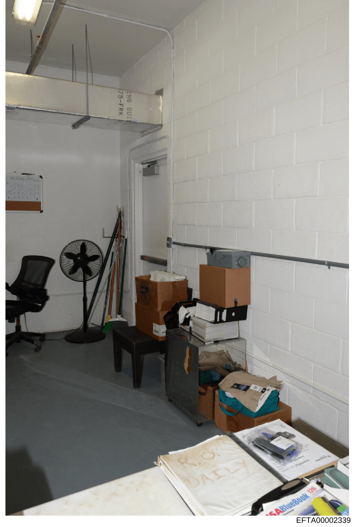 This is an evidence photograph documenting the interior of a storage room or office space containing various items including boxes, office equipment, and personal belongings. The image appears to be part of the investigative record, showing the physi