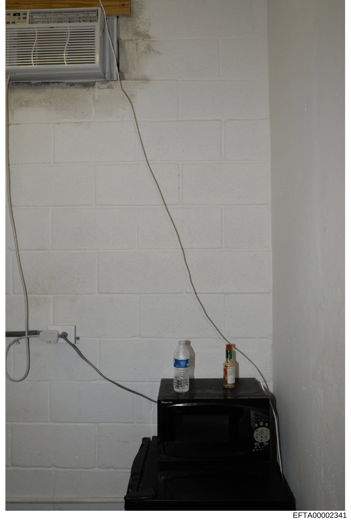 This is an evidence photo showing the interior of a room with a window-mounted air conditioning unit, a microwave oven on a black surface, and various electrical cords. The image appears to be part of the physical evidence documentation from the Epst
