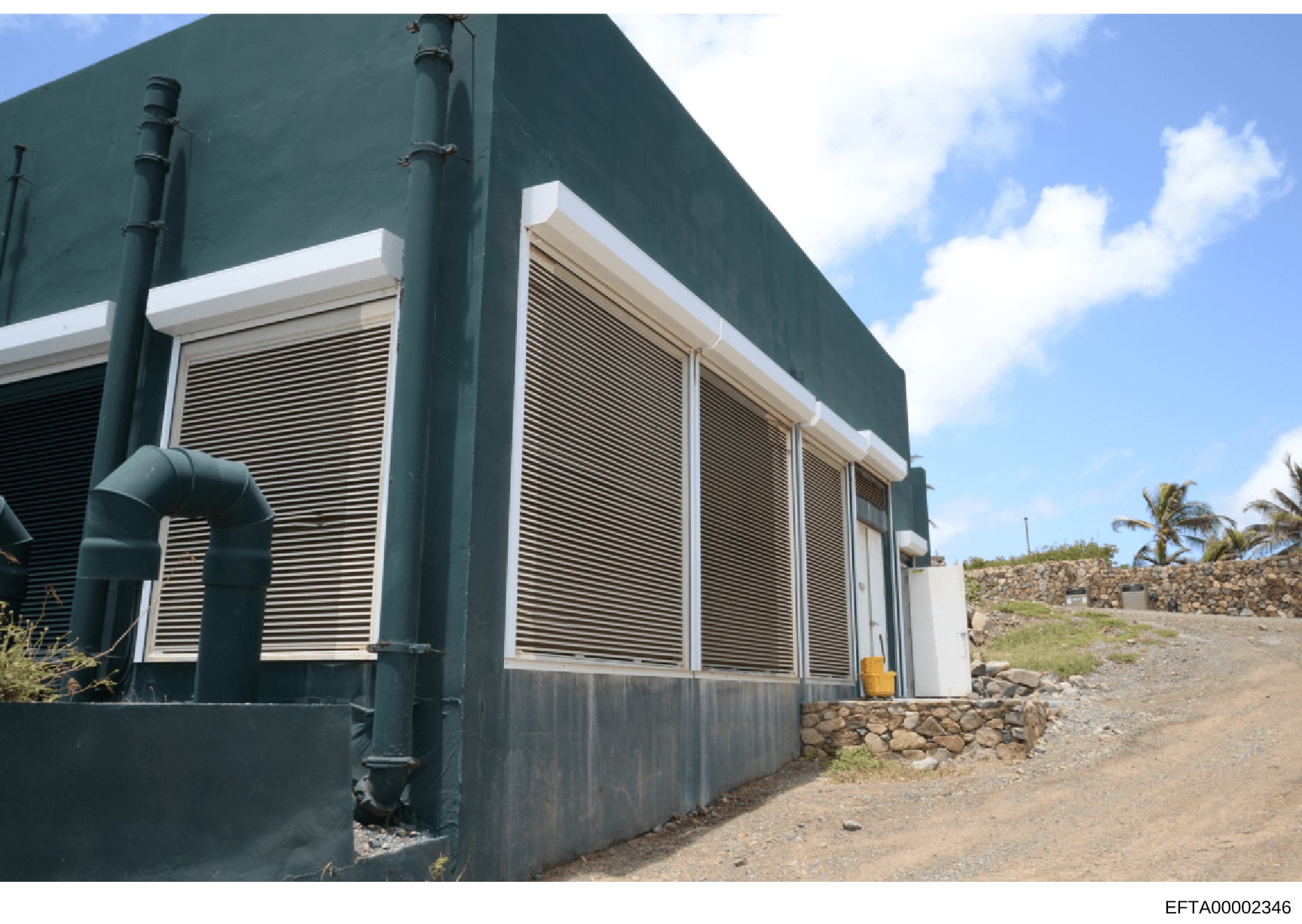 This is a photograph of an industrial or utility building featuring a dark teal/green exterior wall with white-trimmed louvered windows, metal piping, and a gravel/stone foundation. The structure appears to be located in a tropical or semi-arid locat