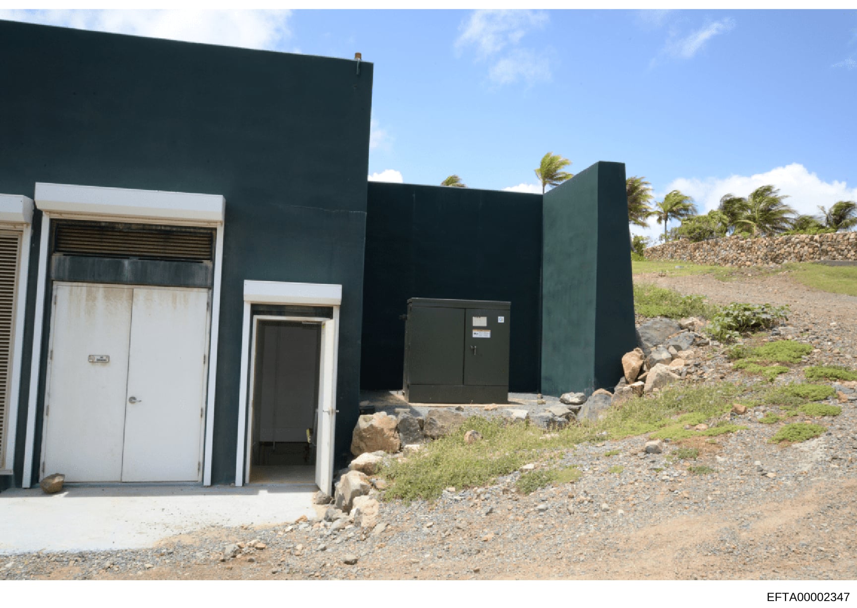This is an evidence photograph showing the exterior of a modern dark-colored building or structure with multiple entrances and utility equipment. The building features white-trimmed doors and a dark metal utility box, situated on a rocky/gravel terra