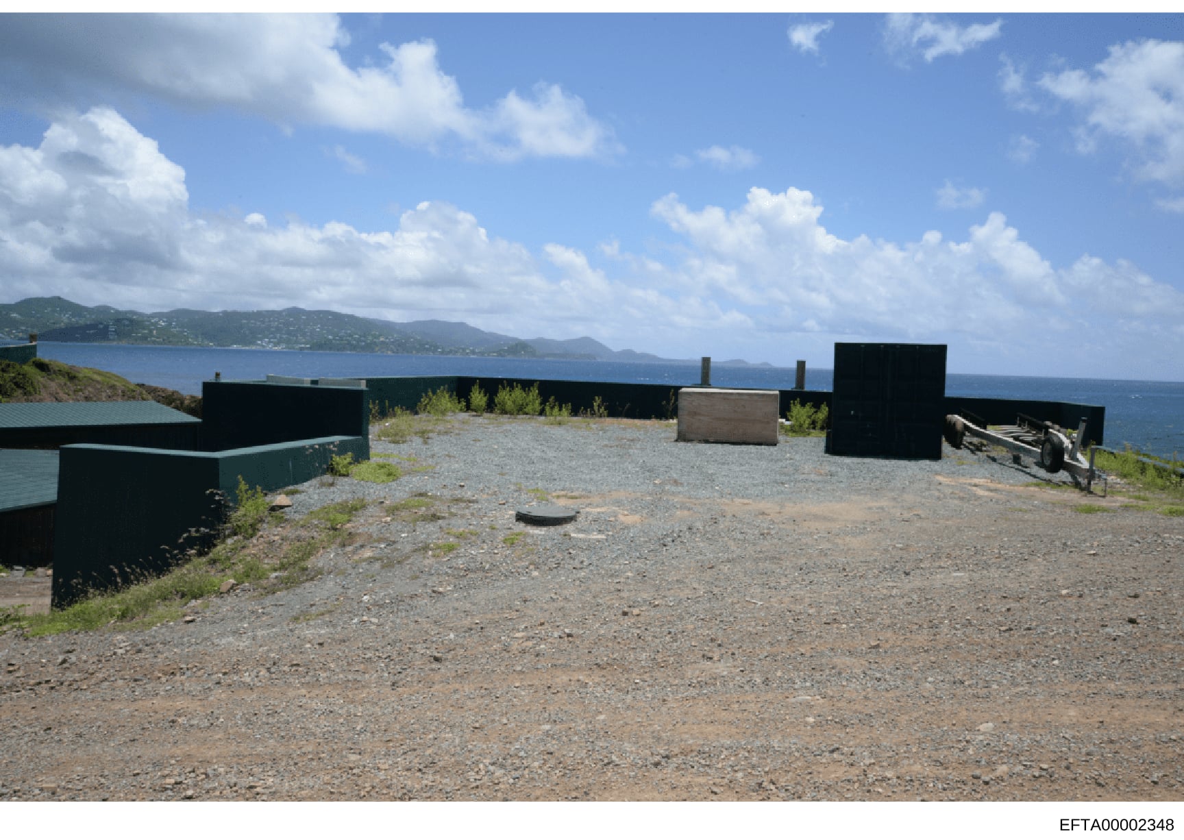 This is a photograph of a waterfront property featuring multiple black-painted structures and storage buildings on a gravel compound overlooking a blue ocean with mountainous islands visible in the distance. The image appears to be evidence documenta