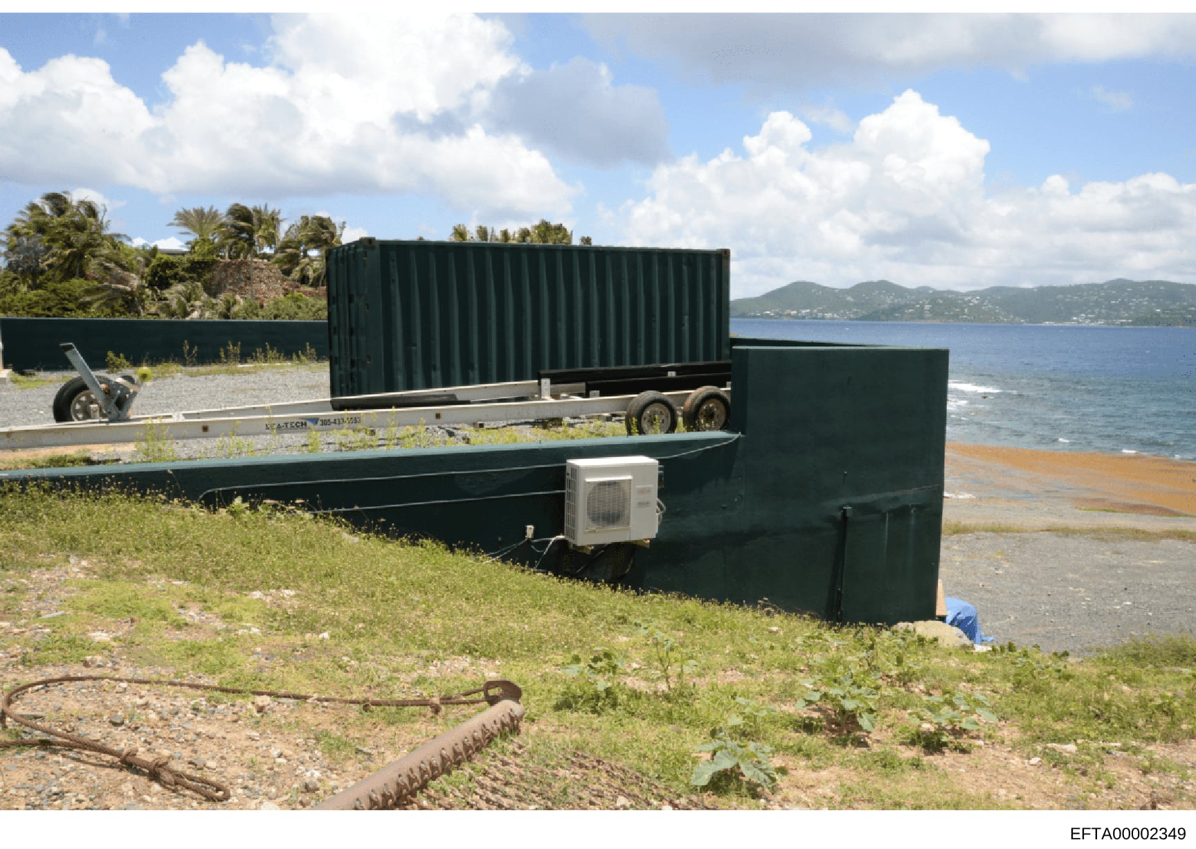 This photograph shows a beachfront property with green metal storage containers and a trailer positioned on sandy/grassy terrain. The image captures what appears to be a coastal residence or property storage area, with ocean and mountainous landscape