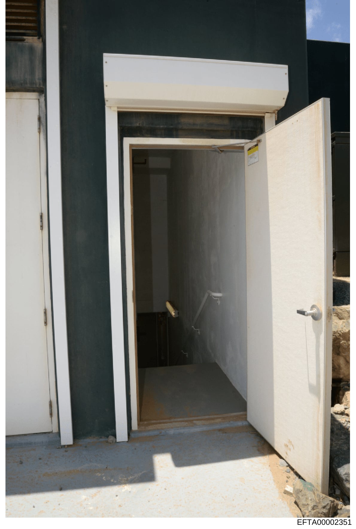 This is an evidence photograph showing the exterior of a white modular or prefabricated building structure with its door open, revealing a dark interior space. The photo appears to be taken outdoors in daylight conditions and includes visible Bates s