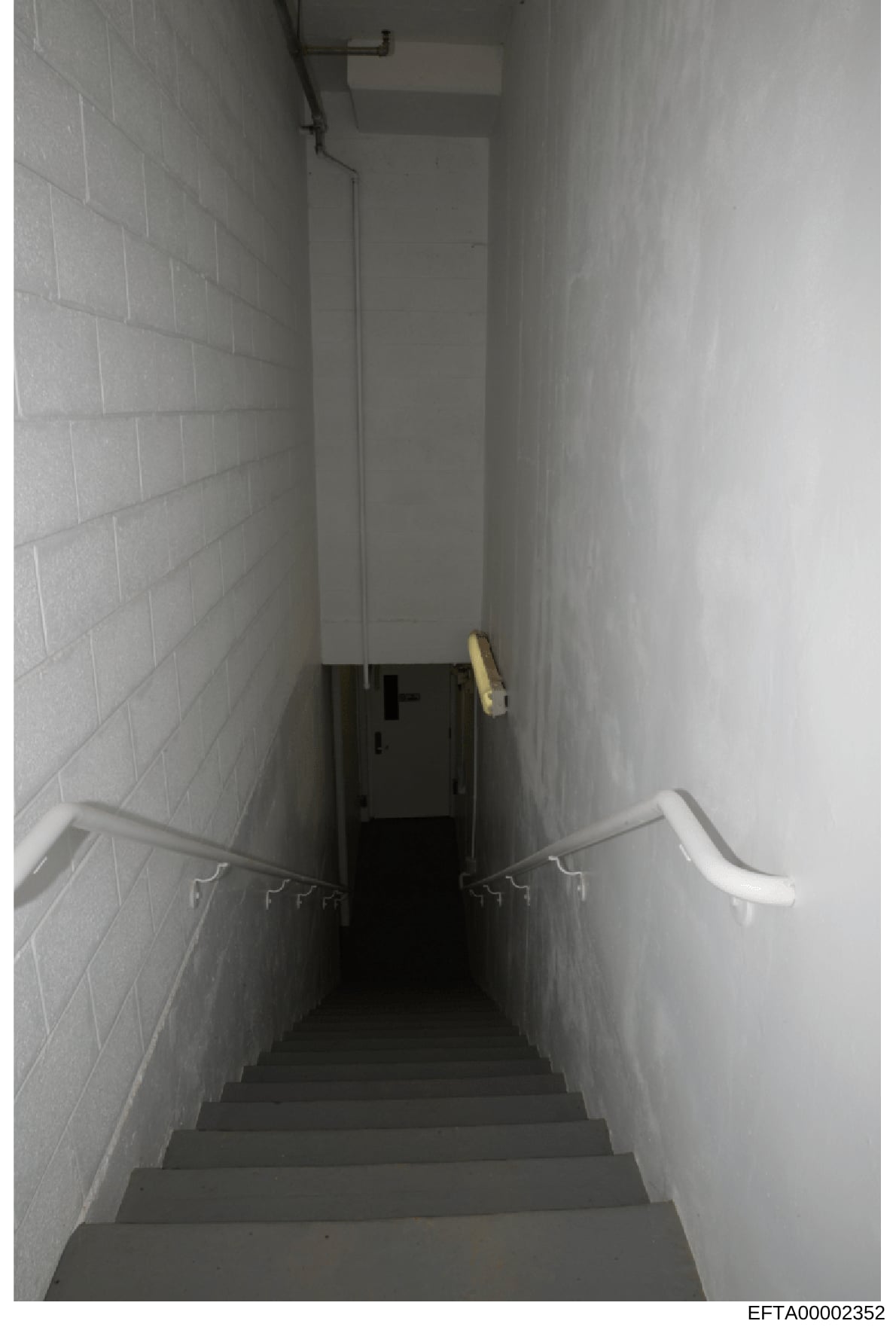 This is a photograph of a narrow, institutional-style stairwell with white cinderblock walls, concrete steps, and minimal lighting. The space appears to be a basement passage or utility area, showing metal handrails and basic infrastructure. The imag