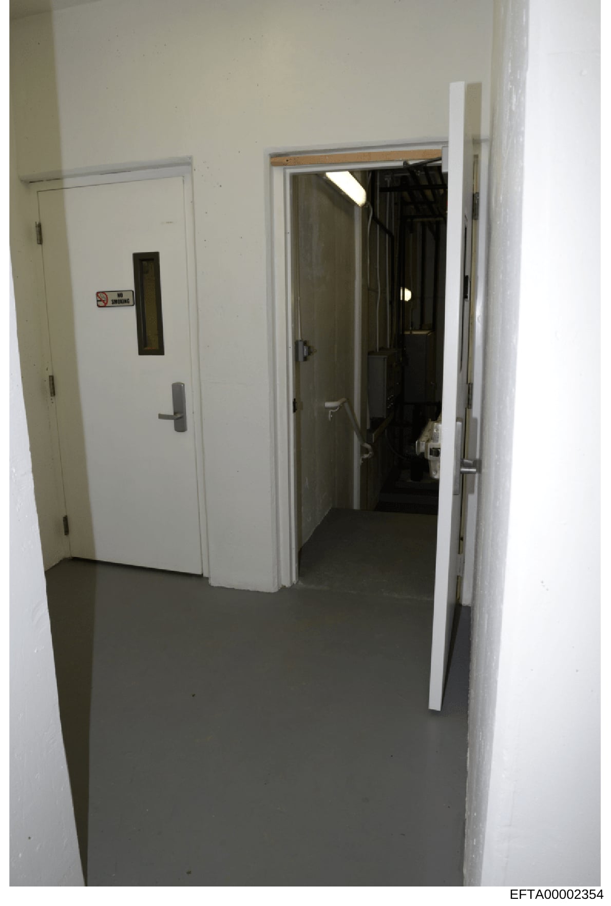 This photograph shows an interior hallway or corridor with white walls and a dark concrete floor. The image features an elevator on the left side marked with a sign, and open doorways leading to other rooms or spaces. The photo appears to be evidence
