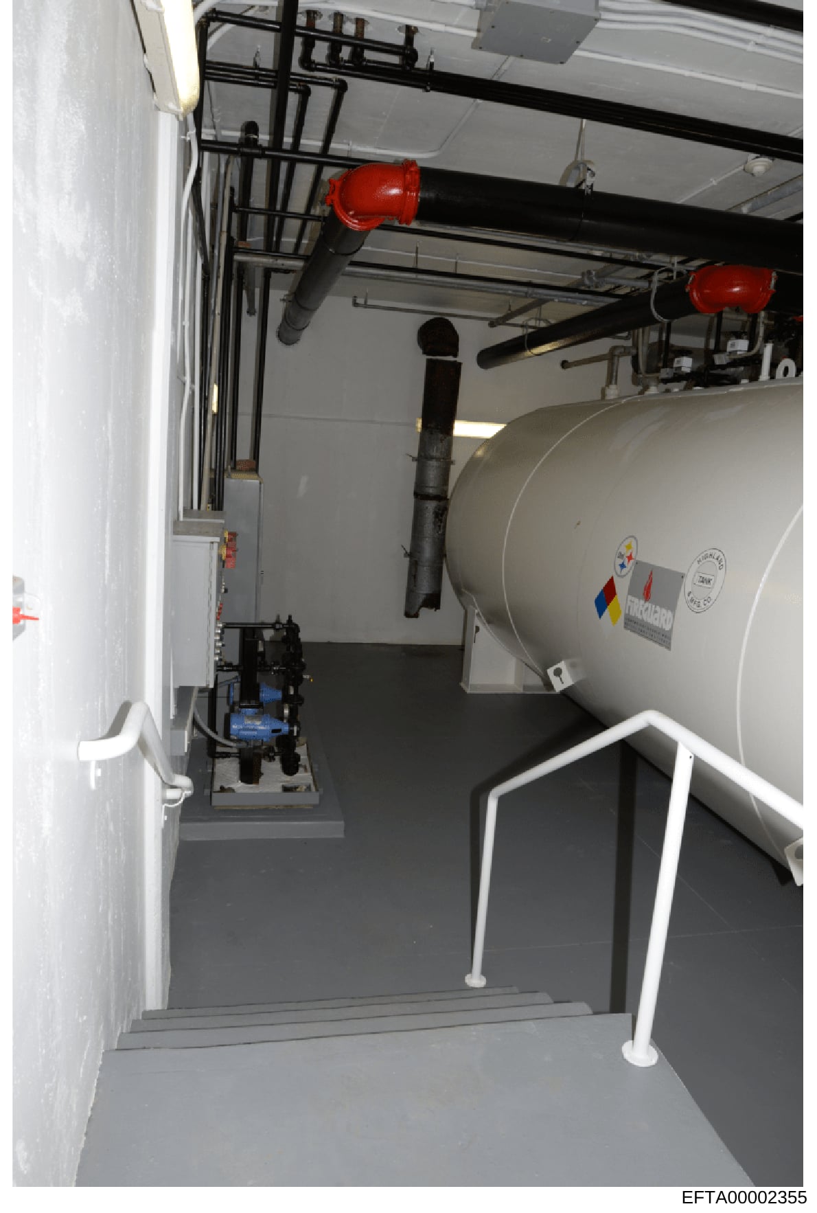 This is a photograph of an interior utility or mechanical room, likely from a residential or commercial property, showing a large cylindrical tank system with piping infrastructure, red safety equipment, and control mechanisms. The image appears to b