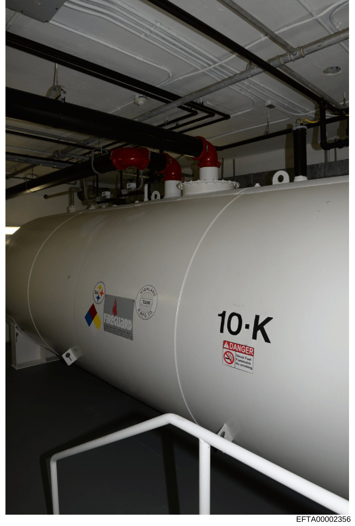 This is a photograph of a commercial fuel storage tank inside an industrial building or facility. The tank is labeled '10-K' and displays warning labels for diesel fuel and flammable materials. The image shows the tank's exterior, overhead piping inf