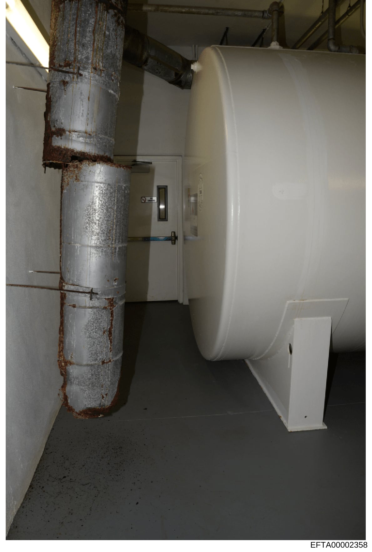 This photograph shows an interior basement or utility room space containing a large white cylindrical storage tank (likely propane or fuel storage) and a heavily corroded metal pipe running vertically along the left wall. The image appears to be evid