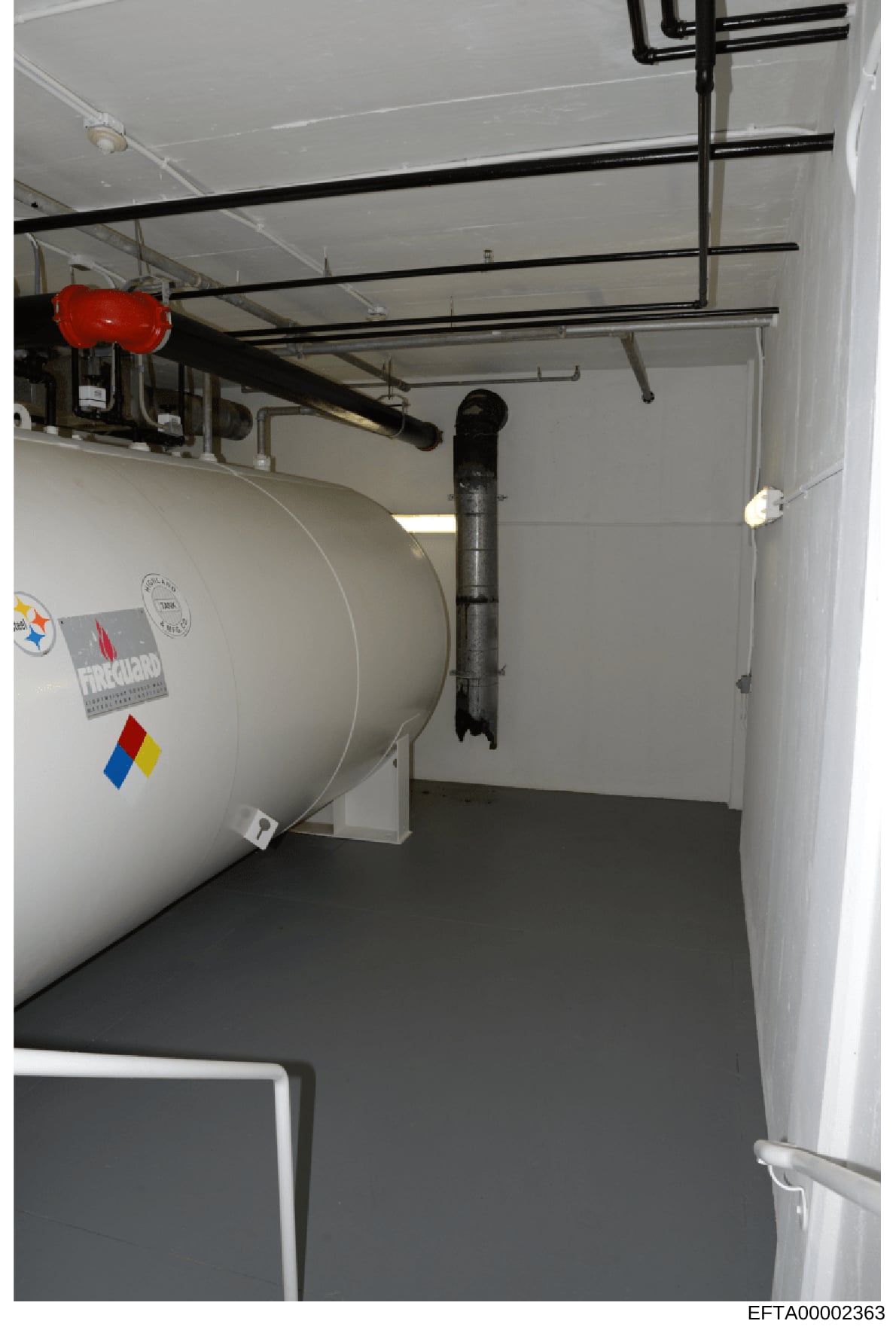 This is an evidence photograph showing the interior of a utility or mechanical room, featuring a large cylindrical heating/storage tank with hazard labeling, black support beams, electrical conduits, and a red emergency device mounted on the ceiling.