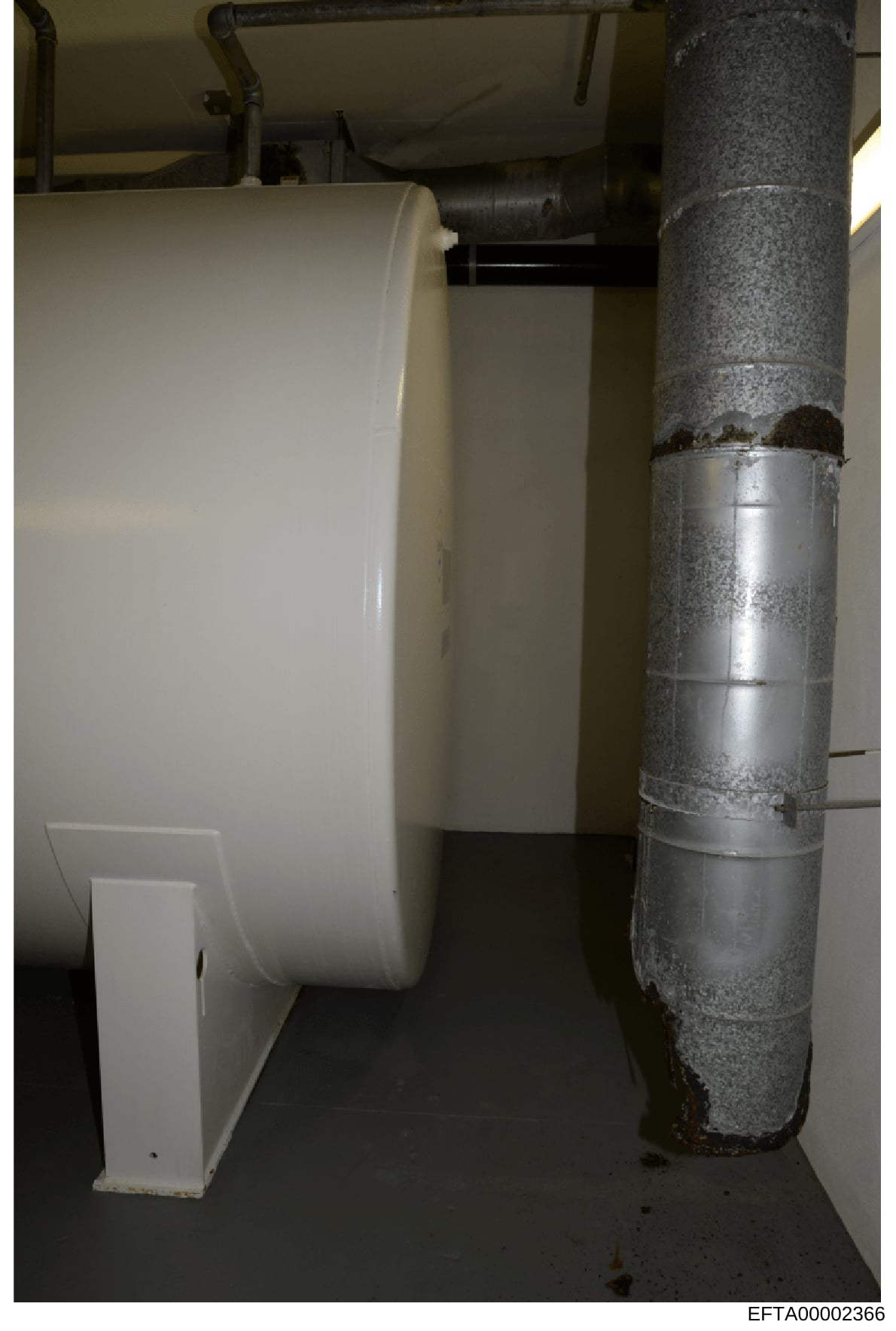This photograph shows the interior plumbing and heating/ventilation infrastructure of a residential space, featuring a white cylindrical tank or container on the left and a large metal ductwork pipe on the right with visible rust and deterioration. T