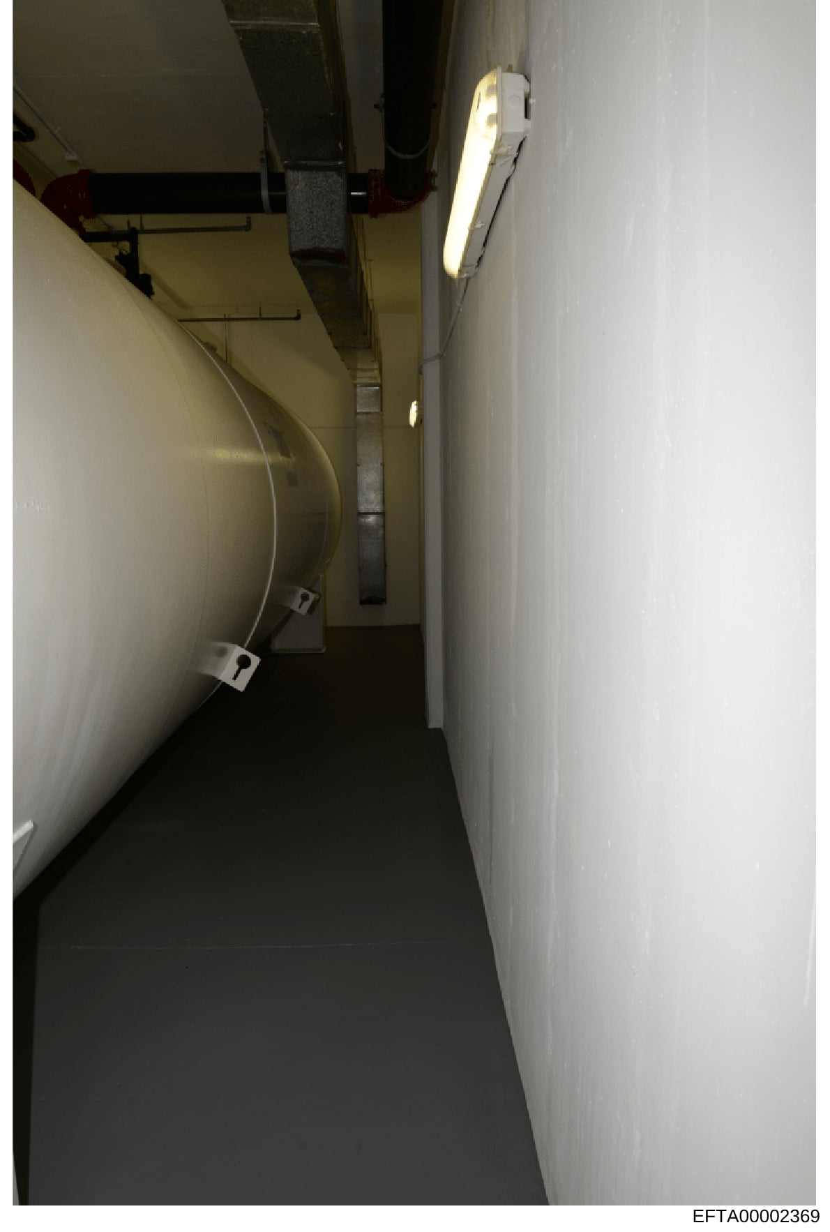 This is an evidence photograph showing a narrow interior hallway or corridor with white walls, fluorescent lighting, and visible mechanical infrastructure including pipes and conduit work. The image appears to be documentation of a building interior,