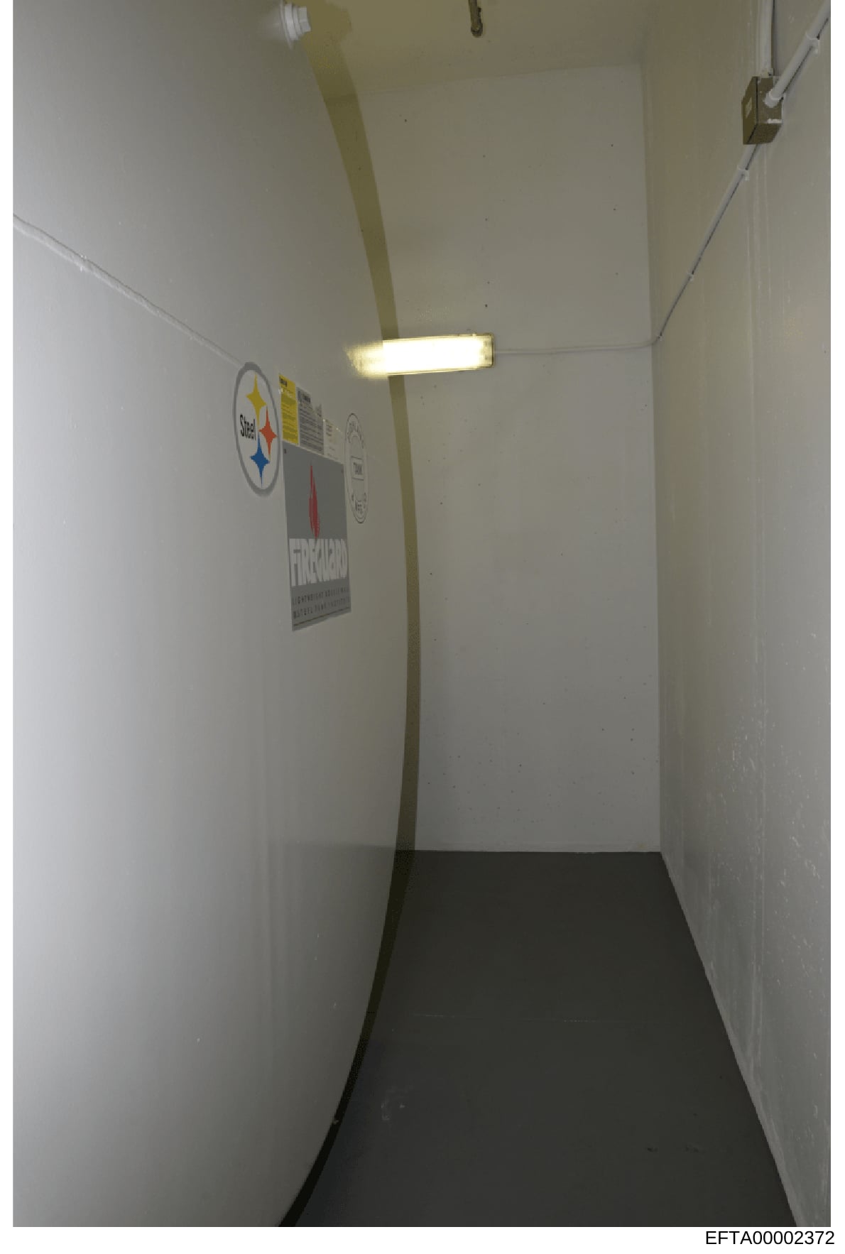This is a photograph of a narrow interior hallway or corridor with white painted walls, a dark floor, and fluorescent ceiling lighting. The space contains two visible posters or signs on the wall, including what appears to be a Pittsburgh Steelers lo