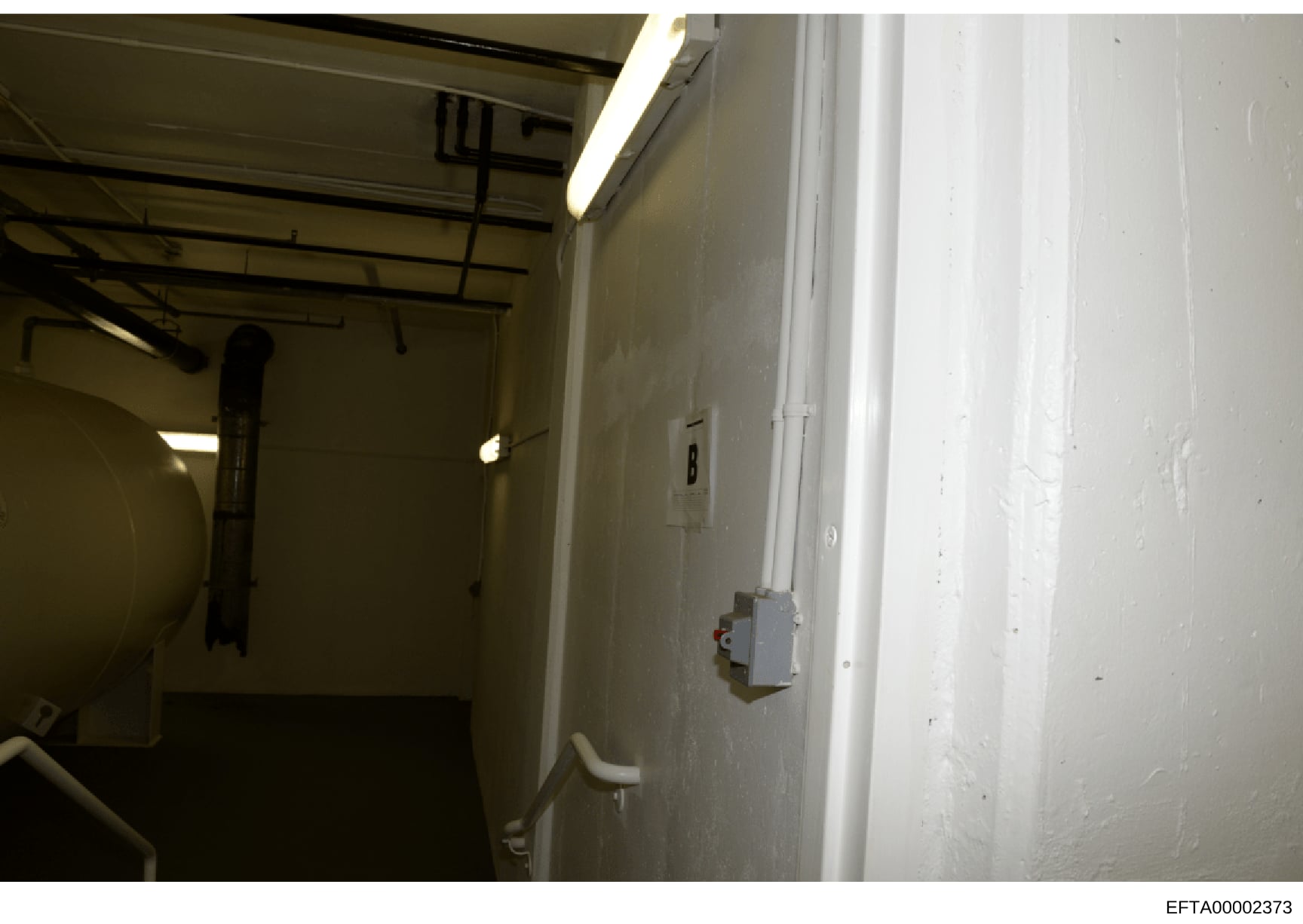 This photograph shows an interior room or utility space, likely part of a residential or commercial property. The space appears largely empty with exposed ceiling beams, fluorescent lighting, and various mechanical/electrical fixtures. The image may 