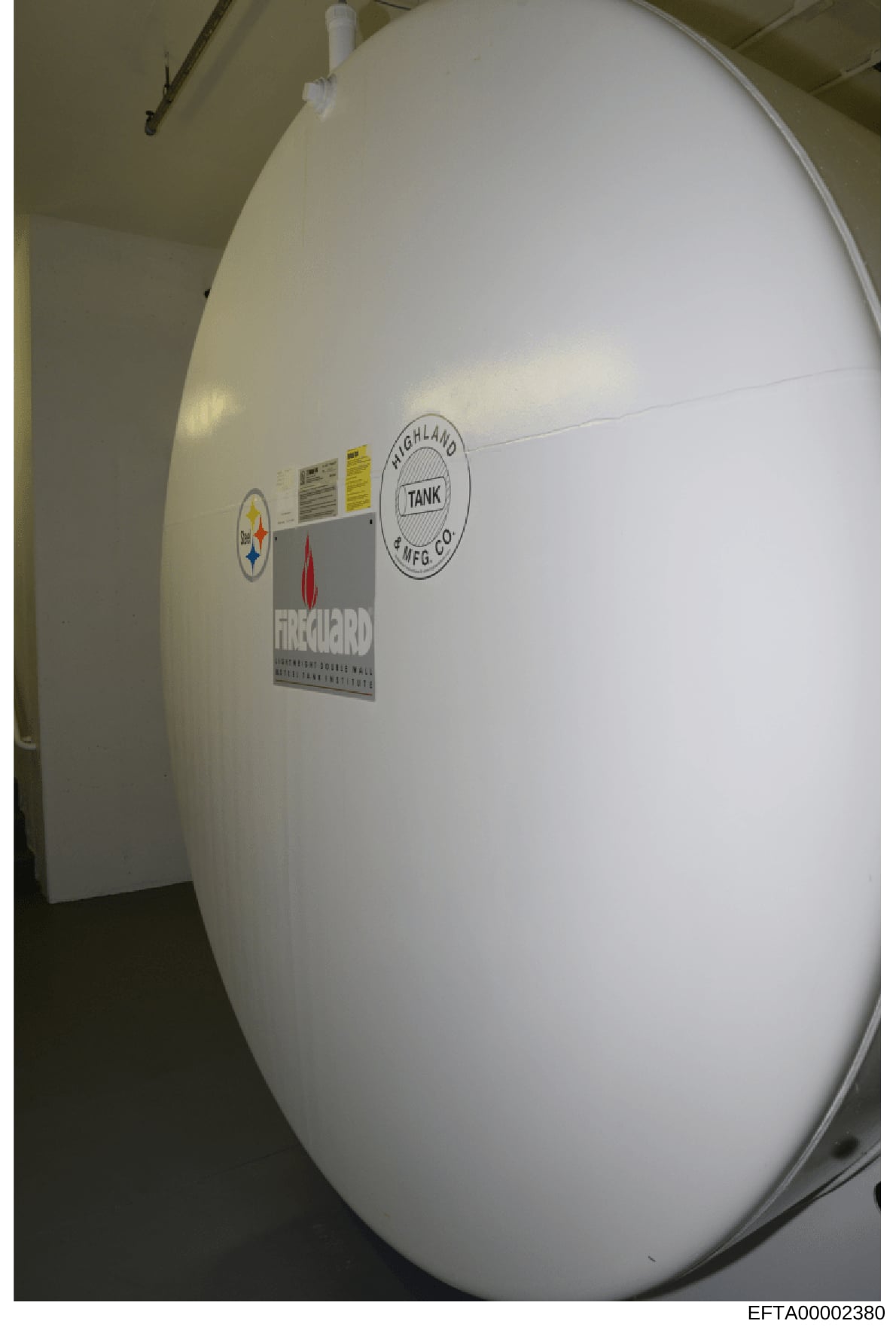 This photograph shows a large white cylindrical storage tank, likely used for water or chemical storage, located indoors. The tank bears manufacturer labels from Highland Tank Manufacturing Company and other regulatory or safety markings. The image a