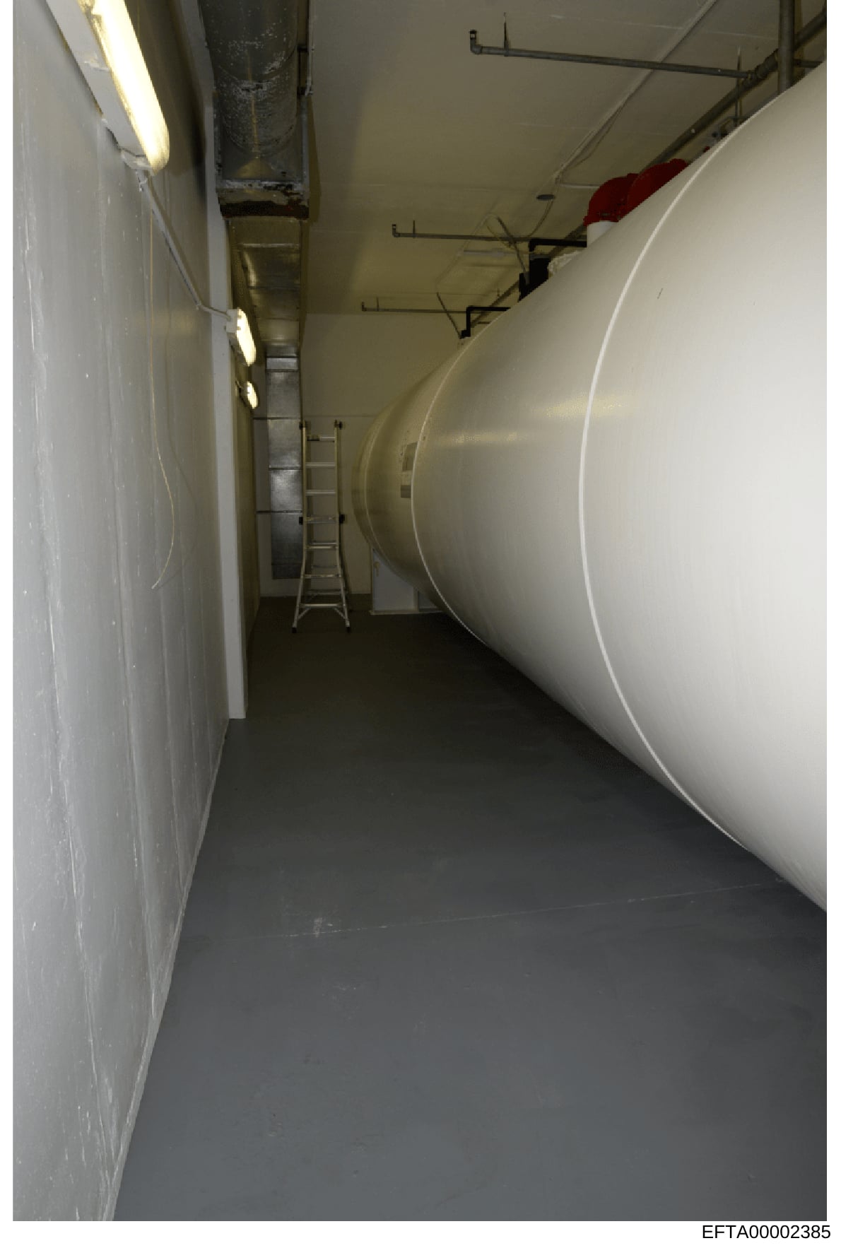 This photograph shows a narrow basement or utility room corridor with a large white cylindrical storage tank running along the right side. The space includes fluorescent lighting, exposed piping, a metal ladder, and what appears to be mechanical infr