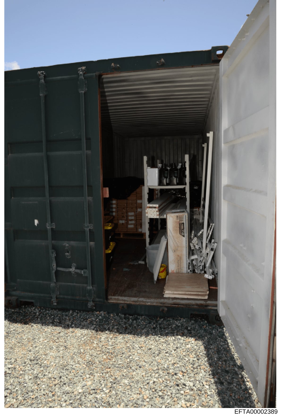 This photograph shows the interior of a large shipping container or storage unit with double doors open, revealing various items stored inside including what appears to be furniture, boxes, wine bottles, and other property. The container shows eviden