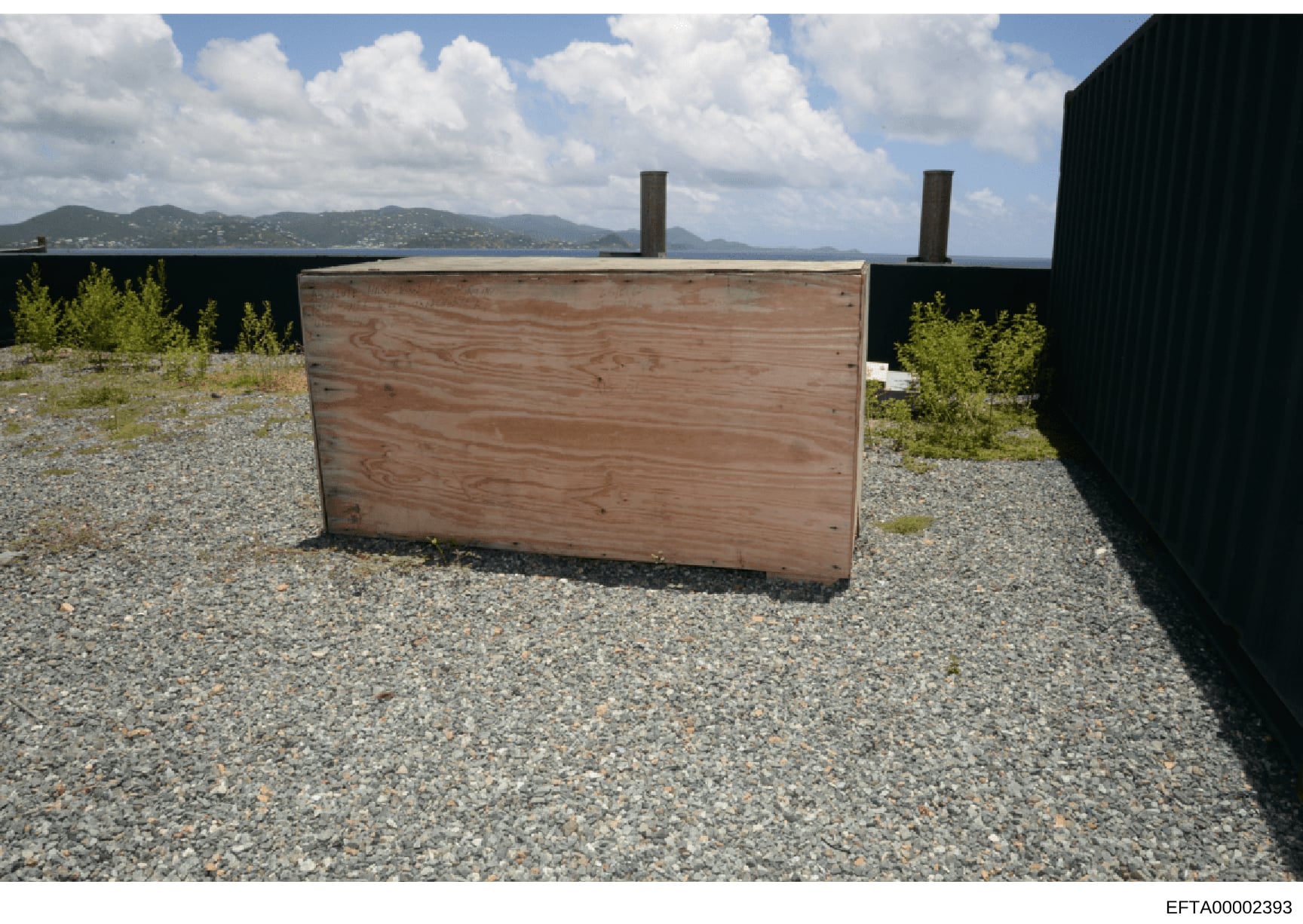 This is a photograph of a wooden storage container or crate positioned on what appears to be a rooftop or elevated platform. The structure features black walls, gravel flooring, and overlooks a tropical island coastline with mountains visible across