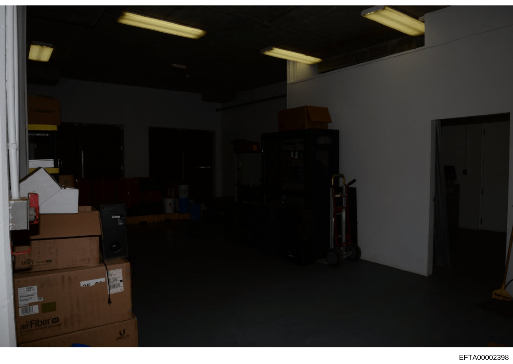 This is a photograph of an interior commercial or warehouse-type space showing a large empty room with fluorescent ceiling lights, white walls, and dark flooring. The room contains several cardboard boxes (including one labeled with 'Fiber' branding)