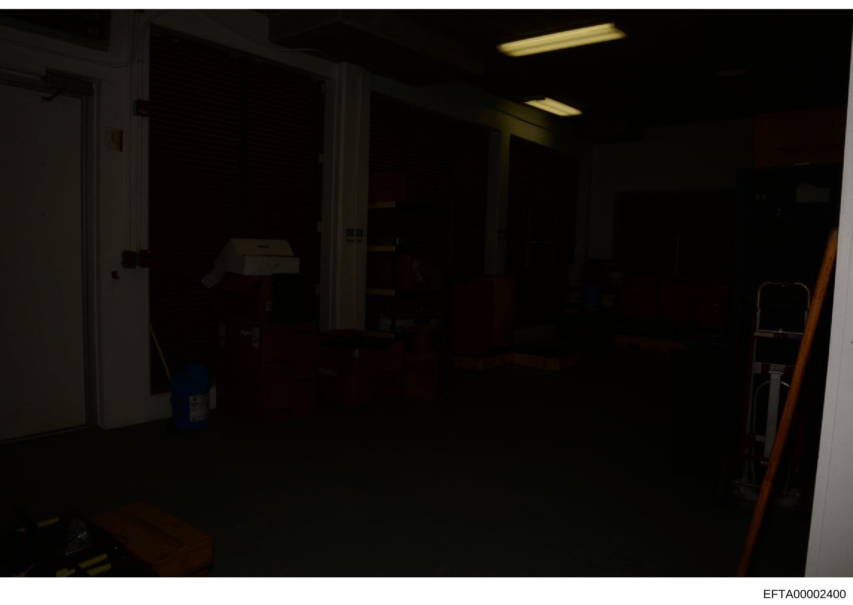 This photograph shows the interior of a dark, sparsely furnished room, likely part of a property being documented during the investigation. The image captures what appears to be a utility or storage area with minimal visible furnishings and equipment