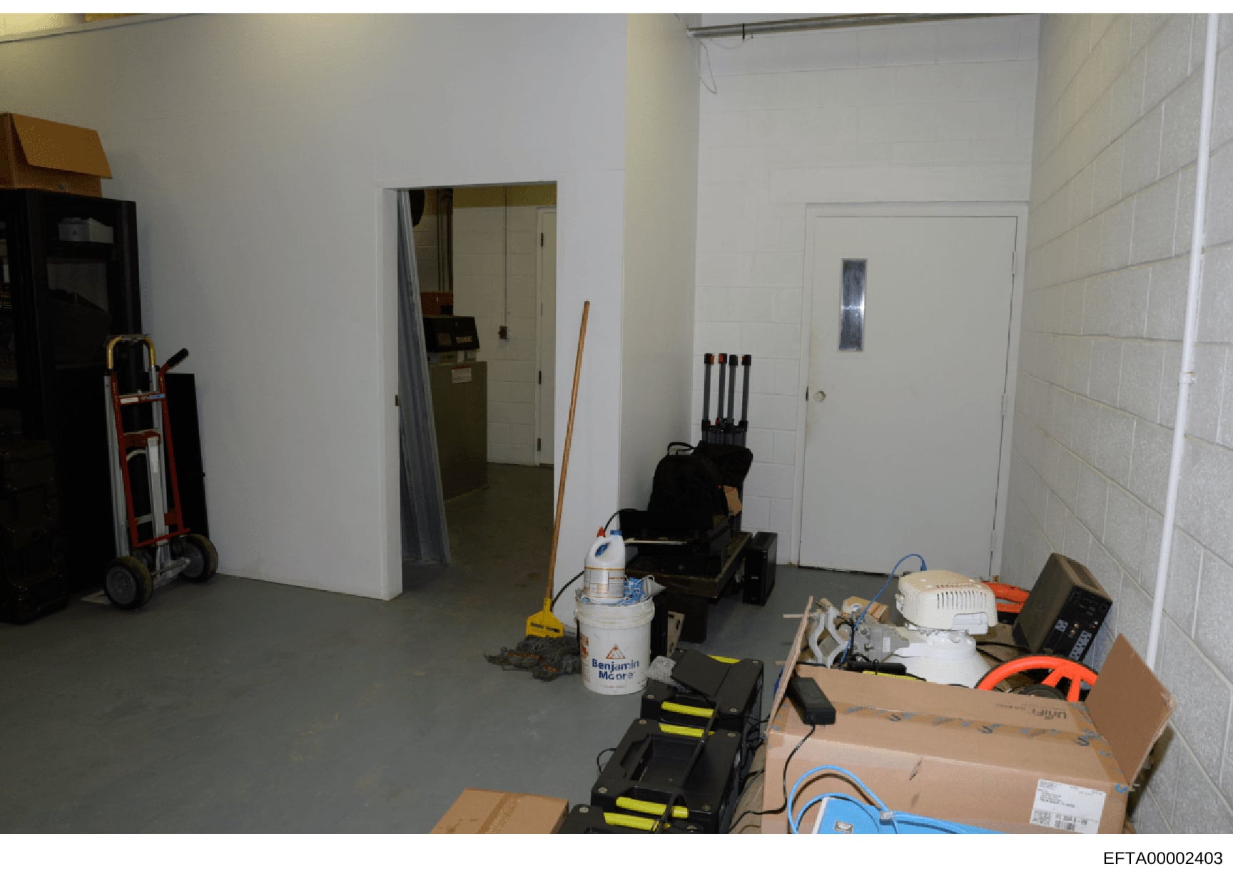 This is an evidence photograph showing the interior of a basement or utility storage area containing various maintenance equipment, tools, and supplies. The image captures cardboard boxes, a hand truck, cleaning supplies (including a Benjamin Moore p