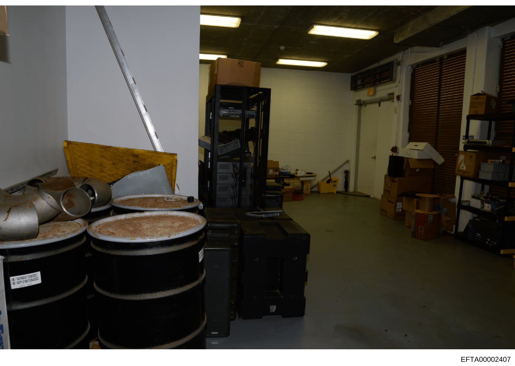 This is a photograph of an interior storage or warehouse facility containing large black drums with lids, metal containers, and various equipment including a black server/equipment cabinet, wooden crates, and fluorescent lighting. The space appears t
