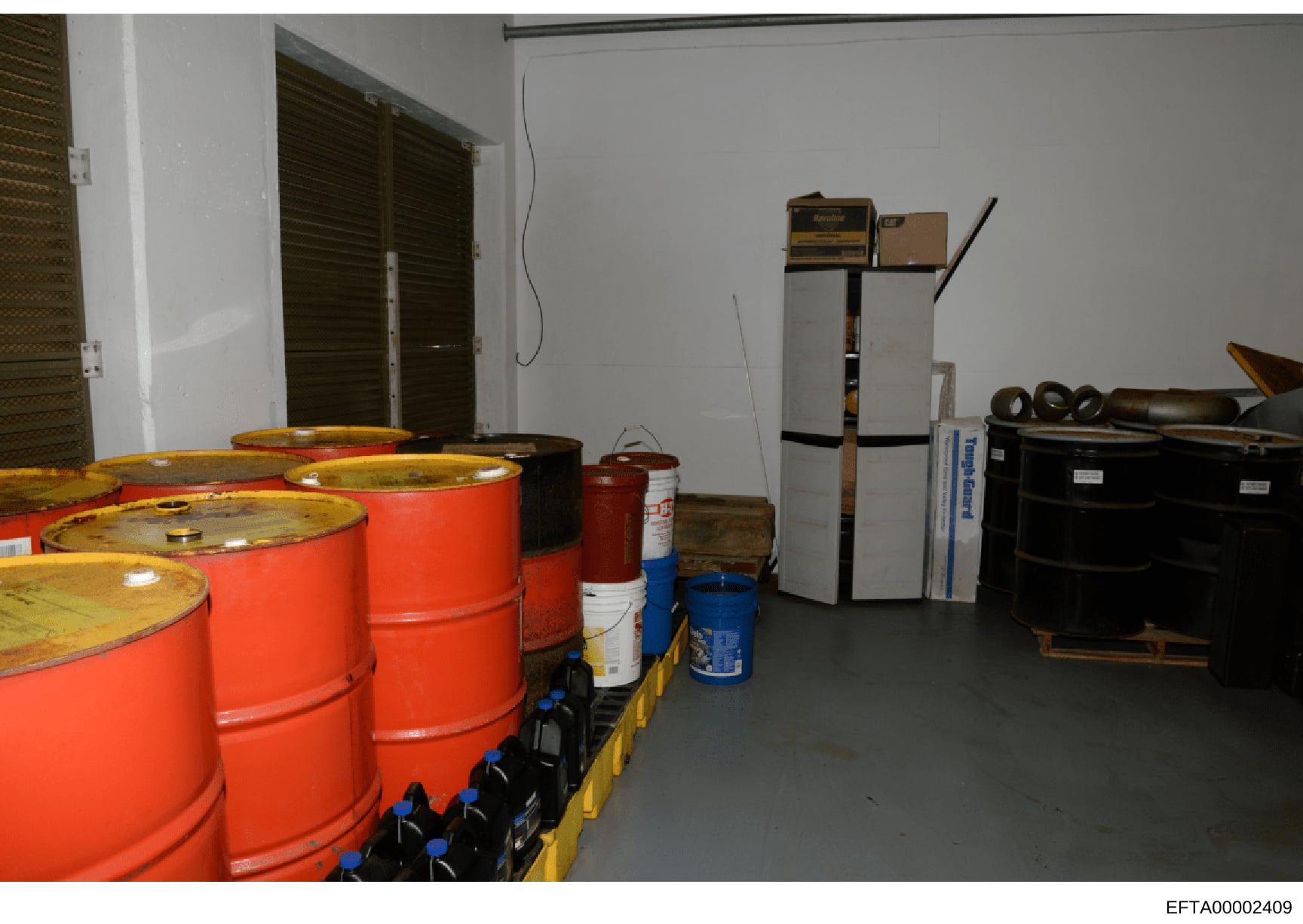 This is an evidence photograph showing a storage or utility room containing multiple large orange industrial drums, various containers, and equipment. The room appears to be part of a property investigation, with items stored on pallets and shelving.