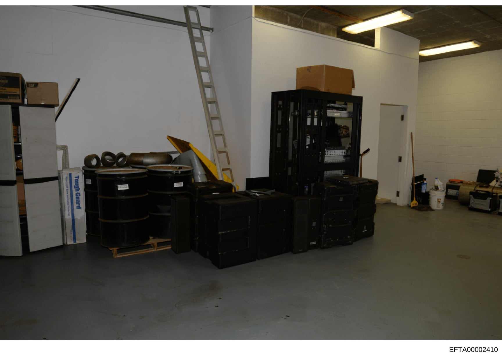 This is an evidence photograph depicting the interior of a storage or warehouse space containing numerous seized items including black storage containers, furniture, equipment, and various household items. The image appears to be part of the forensic