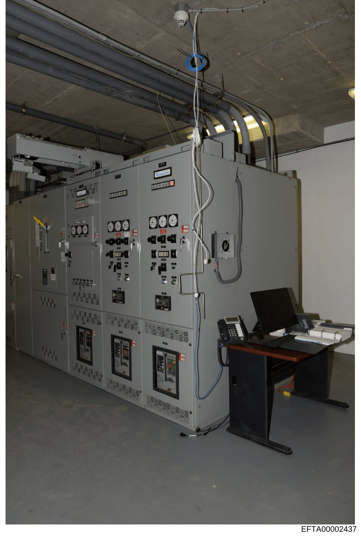 This is a photograph of an industrial electrical control room or equipment facility showing large gray electrical distribution panels, monitoring systems, control consoles, and overhead wiring infrastructure. The image appears to be documentation of 