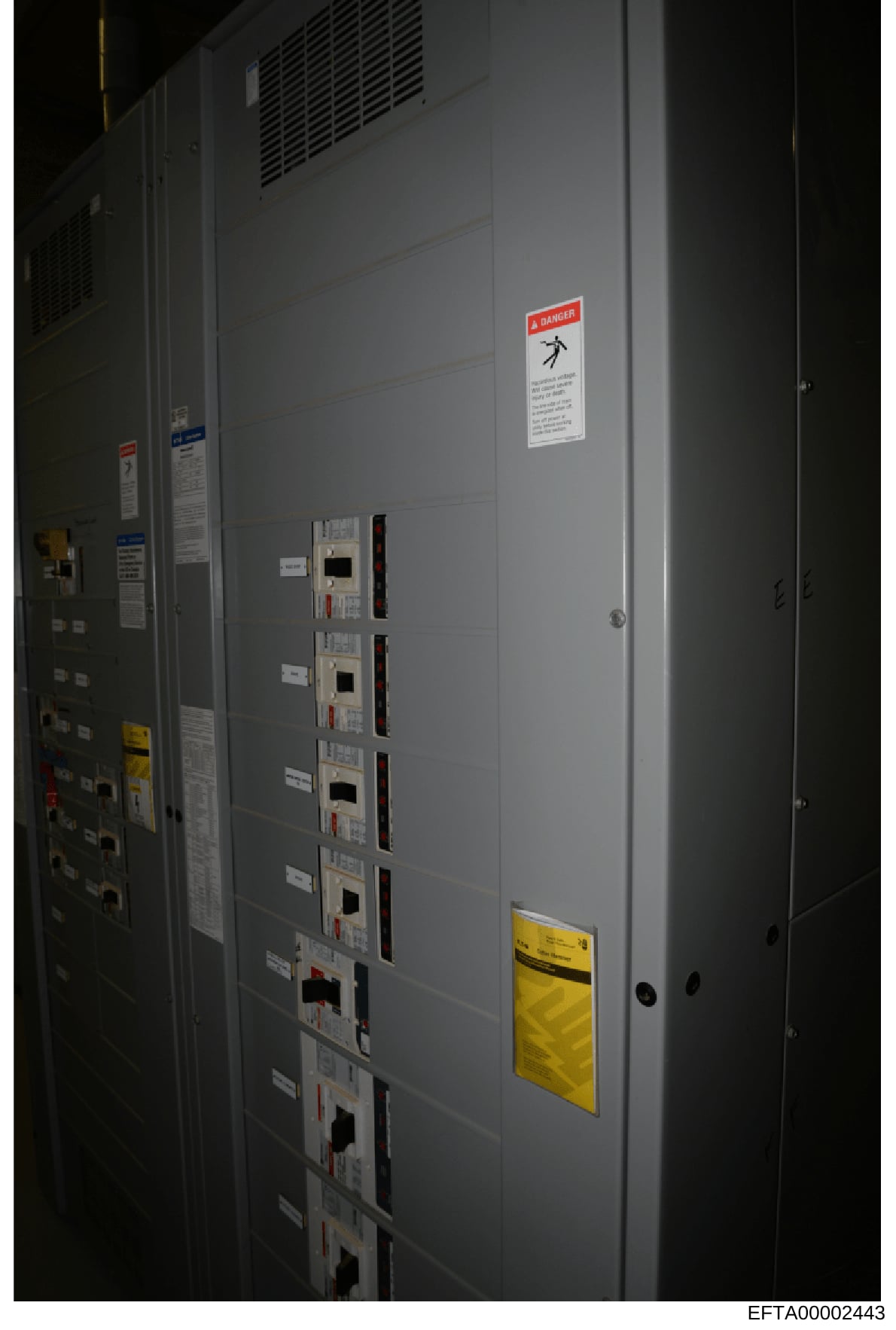 This photograph shows the interior of an electrical equipment room or utility closet, featuring a large gray panel cabinet with multiple circuit breakers or electrical control units, ventilation grilles, and prominent safety warning labels including 