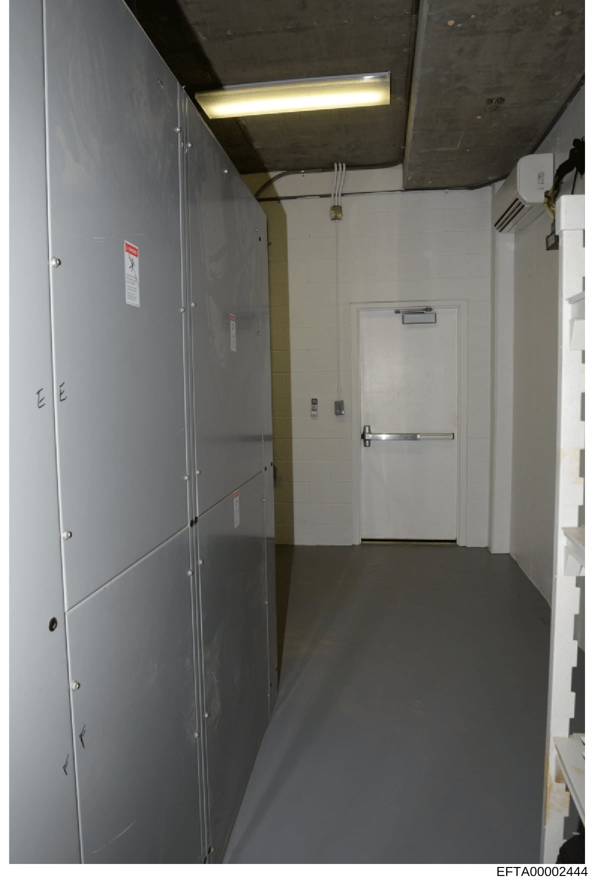 This photograph depicts an institutional or commercial interior hallway, featuring white and gray walls, fluorescent lighting, and multiple doors with safety signage. The space appears to be part of a building facility under investigation, with visib