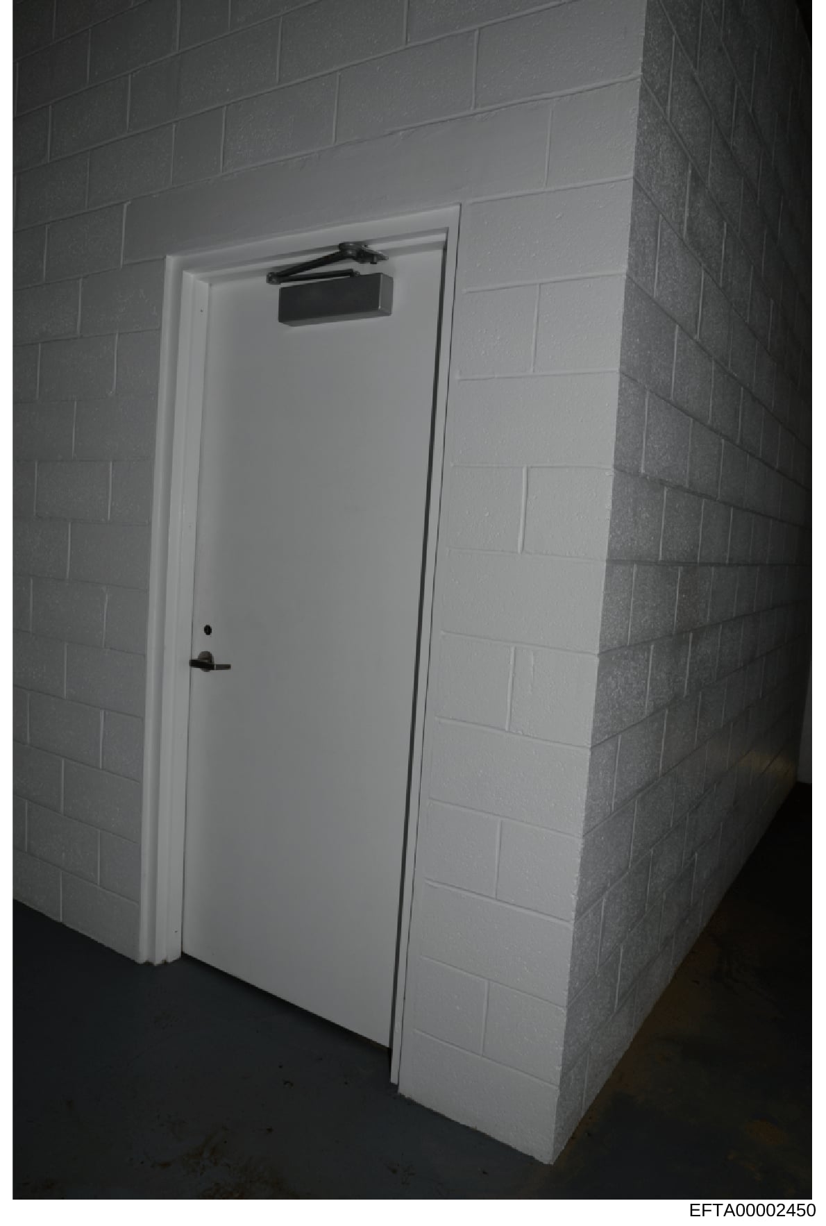 This is a black and white photograph of a white-painted interior door with an automatic closer mounted above the frame. The door appears to be in a basement or institutional setting with concrete block walls. The photo was taken as part of evidence d