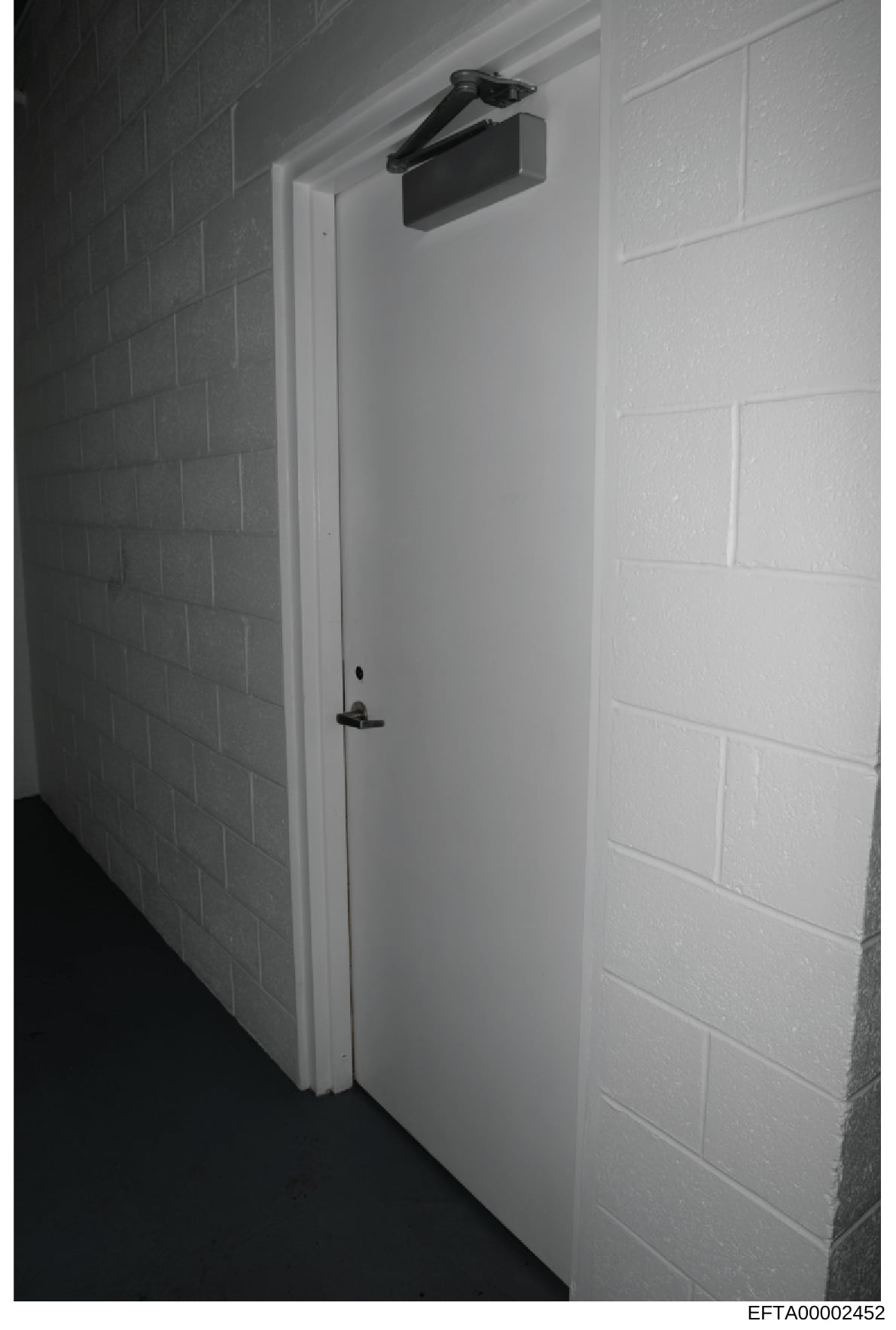 This is a black and white photograph showing an interior doorway with white painted walls and brick detailing. The door features a white frame, black handle hardware, and a black automatic door closer mounted at the top. The image appears to be evide