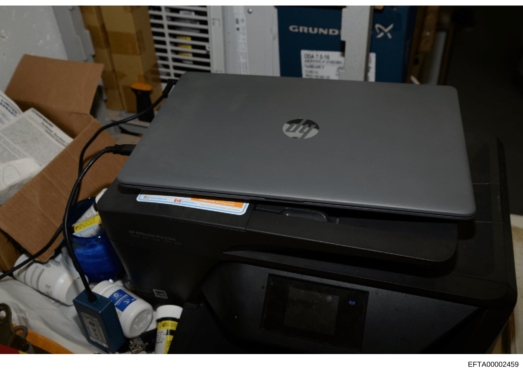 This is a photograph of computer equipment seized as evidence, showing an HP laptop positioned on top of a black printer, with various cables and supplies visible in the background. The image documents the physical state of electronic devices during
