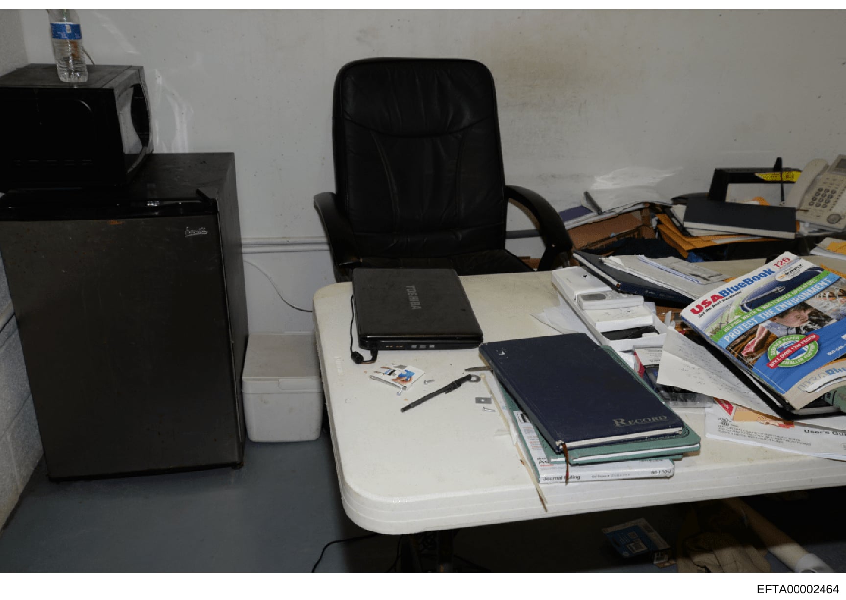 This is an evidence photograph showing an office workspace containing a desk with computer equipment, peripherals, notebooks, magazines, and various papers. The image appears to be from a property search or seizure during the investigation, documenti