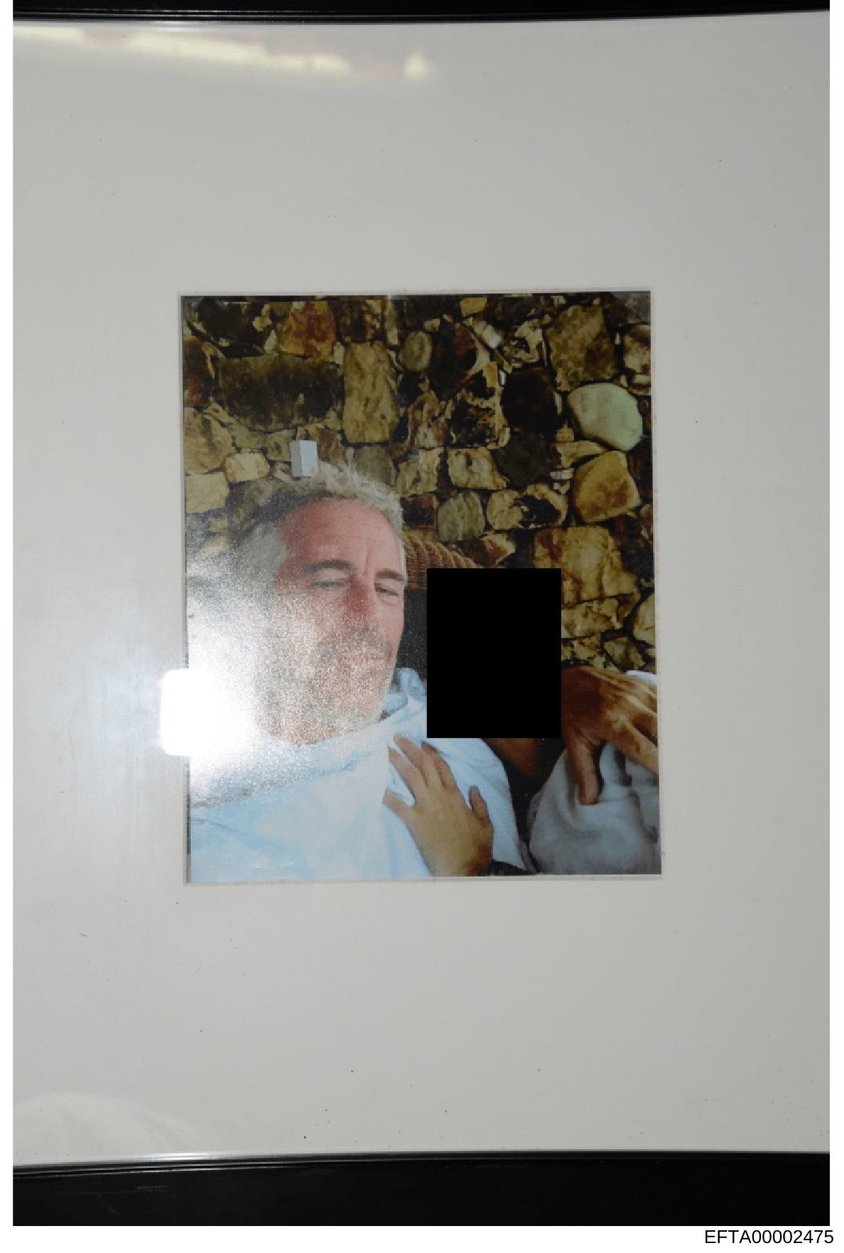 This is an evidentiary photograph from the Epstein investigation files showing a person wearing a light blue shirt against a decorative stone or tile background. A significant portion of the individual's face and body has been redacted with black cen