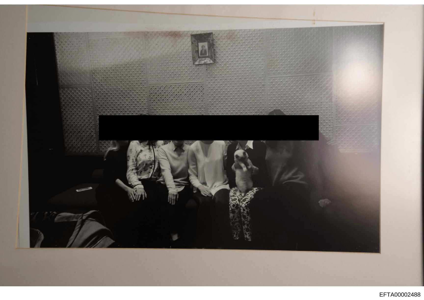 This is a black and white photograph showing four individuals seated together on what appears to be a dark sofa or couch in an interior room with textured wallpaper. A significant portion of the photograph has been redacted with a black bar obscuring
