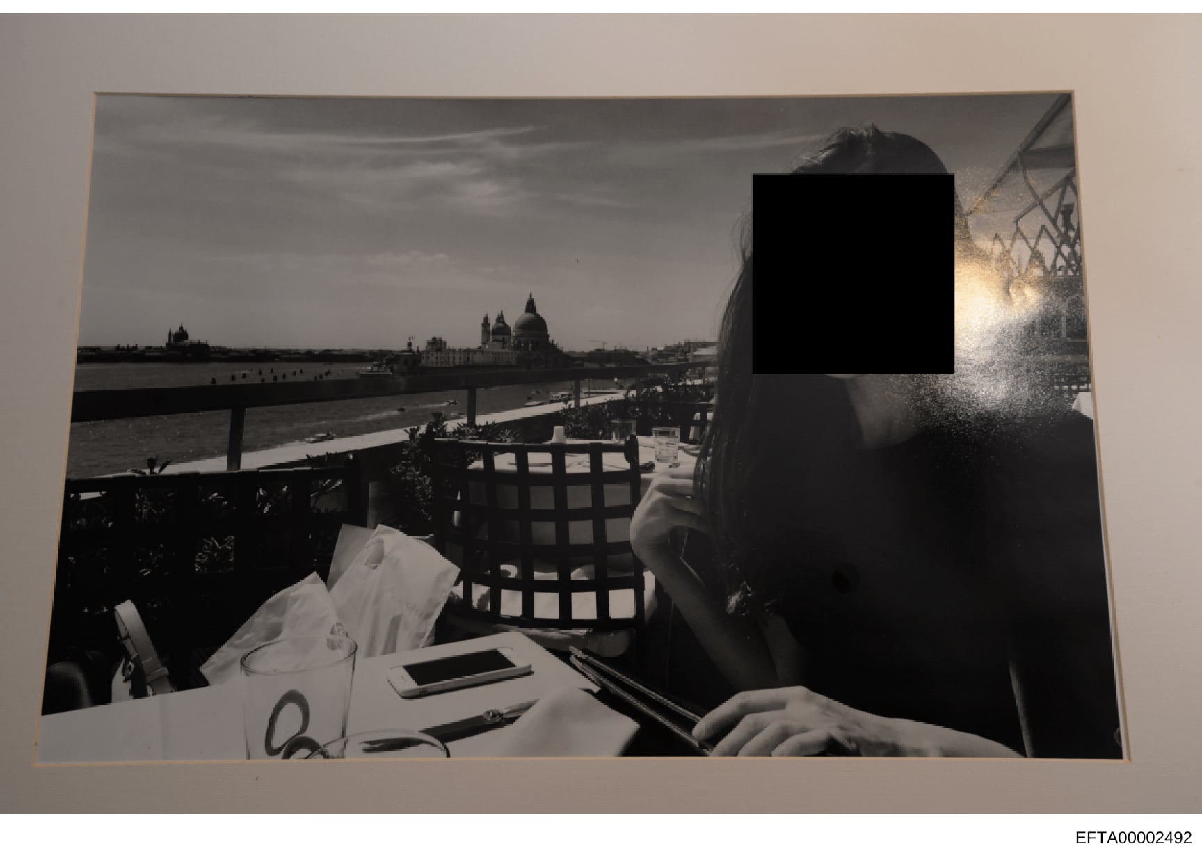 Black and white photograph showing a woman seated at an outdoor waterside dining venue. The image captures a scenic European location with distinctive architecture including domed buildings visible across the water, consistent with Venice, Italy. The
