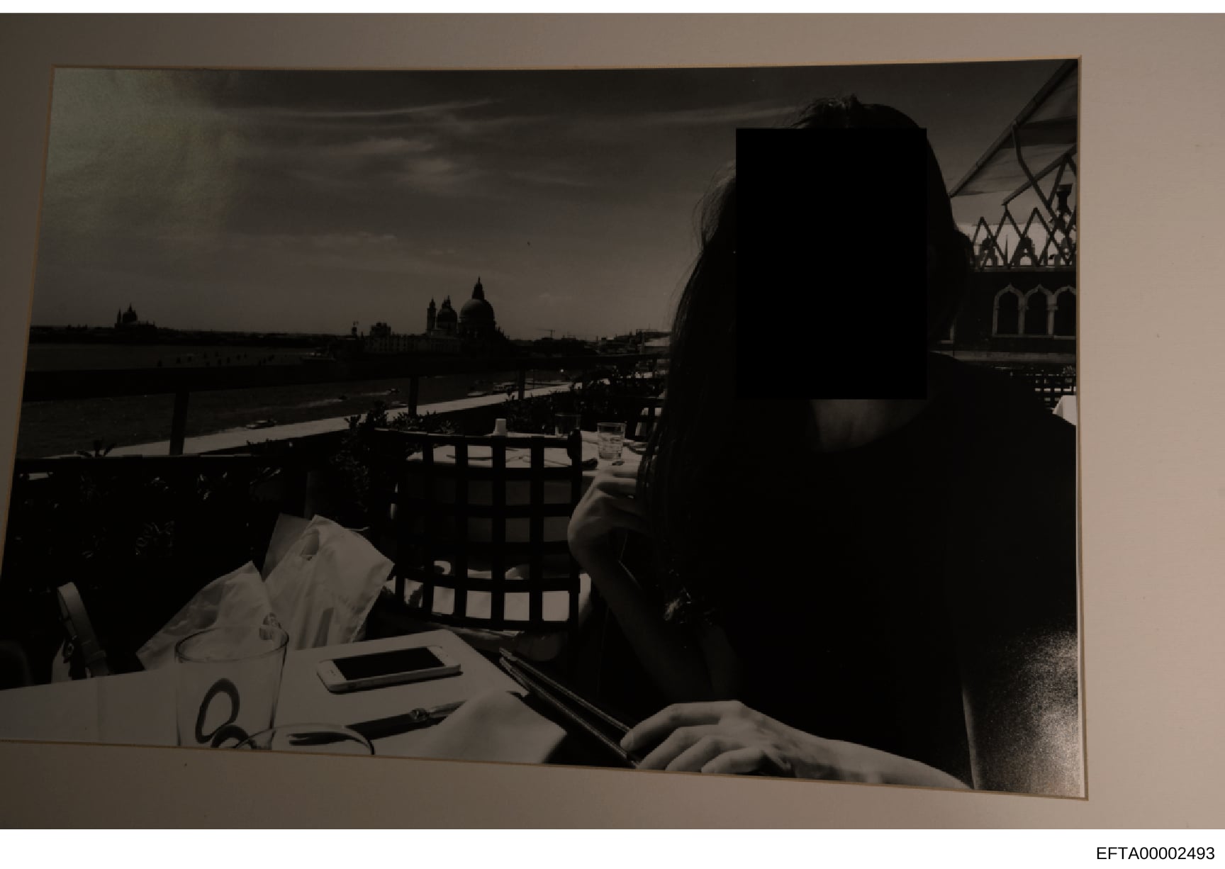 This is a black and white photograph showing a woman seated at an outdoor dining table overlooking a waterway with European architectural landmarks visible in the background. The image appears to be taken at a restaurant or café with a scenic view. T