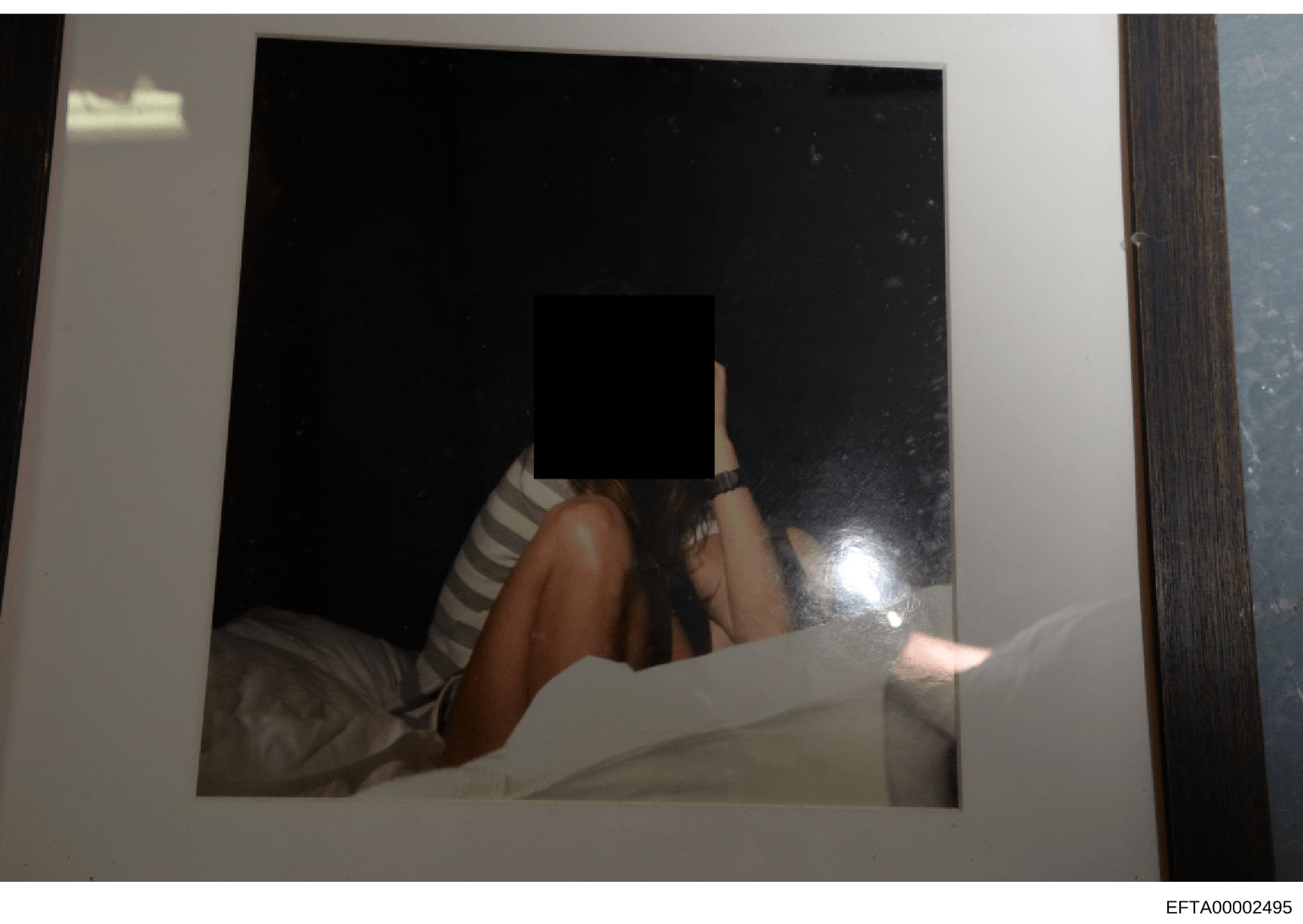 This is an evidence photograph from the Epstein investigation files showing a person with their face obscured by a black square. The image appears to depict an individual on a bed or similar surface wearing striped clothing. The obscuring of the face