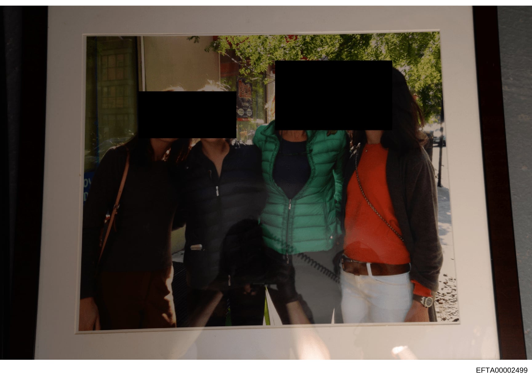 This is an evidence photograph showing three people standing outdoors on a street with trees and storefronts visible in the background. The faces of all three individuals have been redacted with black bars for privacy or investigative purposes. One p
