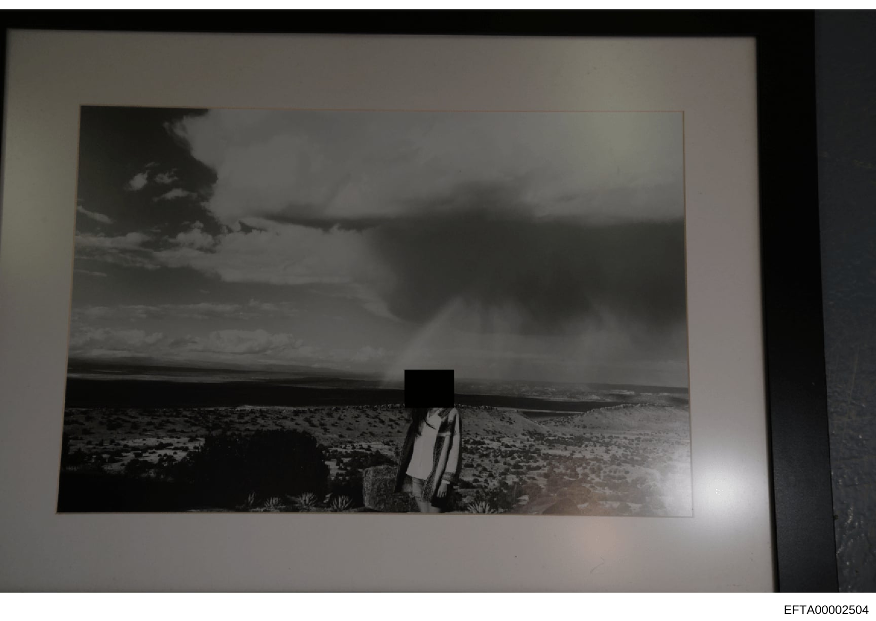 This is a black and white photograph showing a desert landscape with sparse vegetation. A human figure in the center of the image has been redacted with a black box obscuring their identity. The photo appears to be evidence documentation from the inv