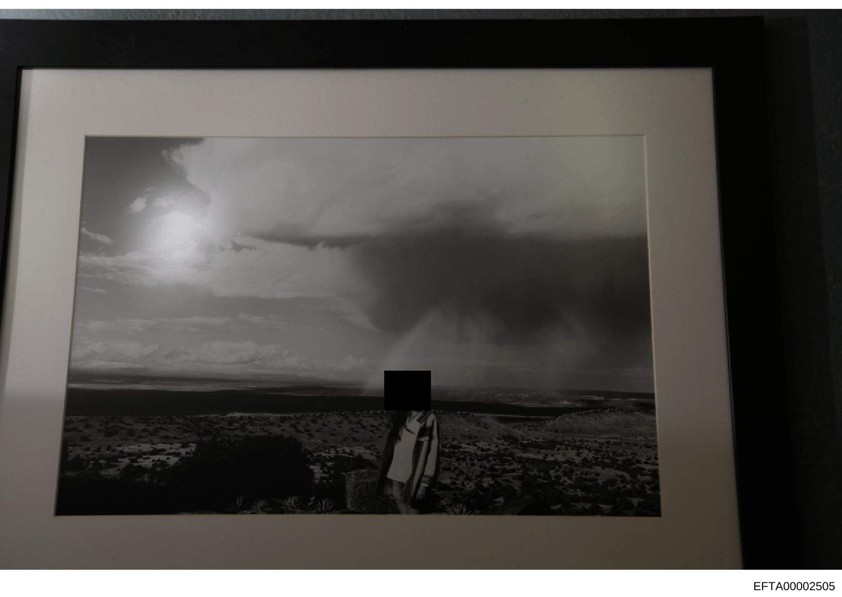 This is a black and white photograph showing a figure standing in an arid, desert landscape with sparse vegetation and flat terrain extending to the horizon. The person's head is redacted with a black box. The photo appears to be evidence documentati