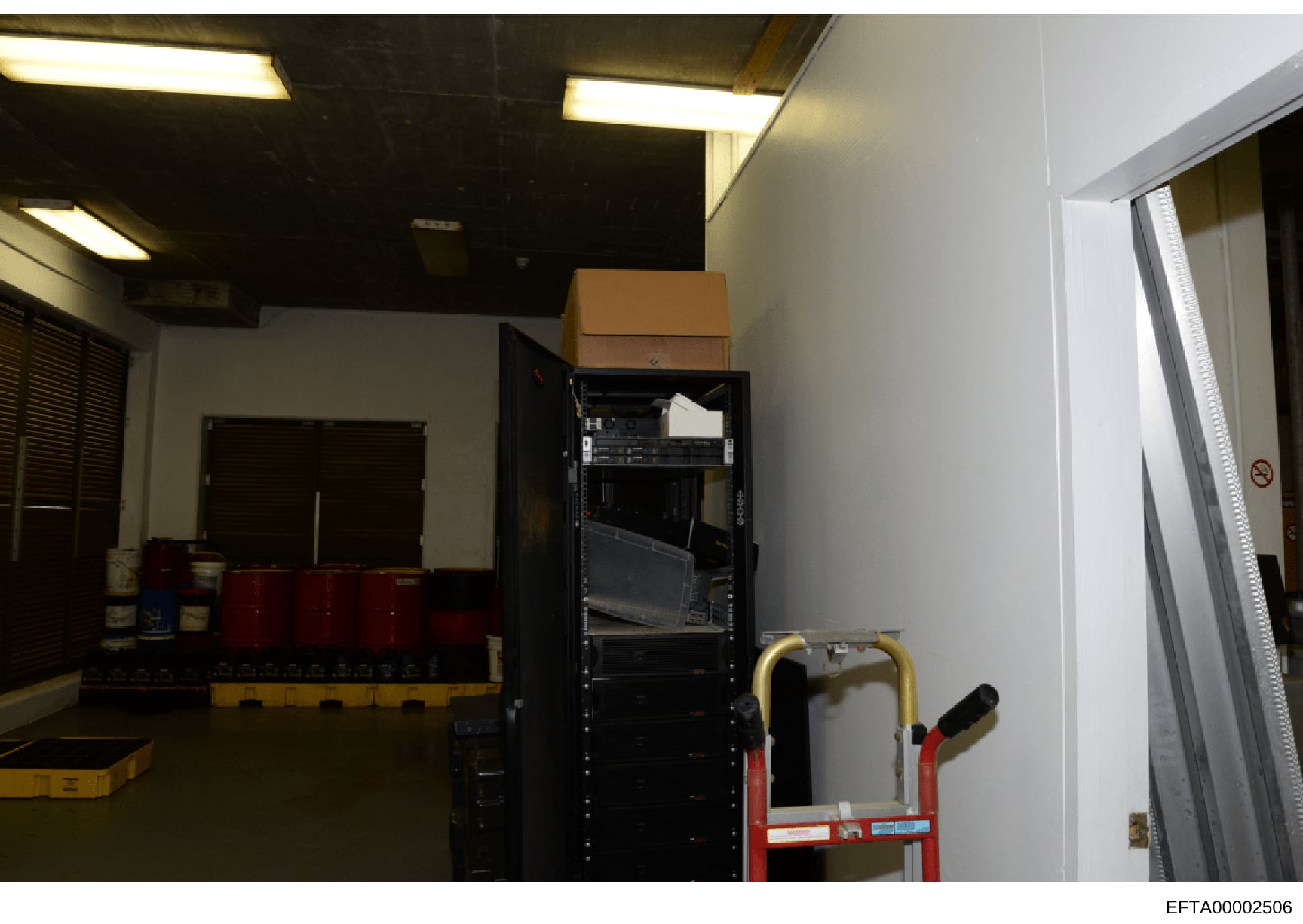 This is an evidence photograph showing an interior storage or equipment room containing a tall black server/equipment rack, red storage containers, a cardboard box, and a red hand truck. The room has fluorescent ceiling lights, white and dark walls,