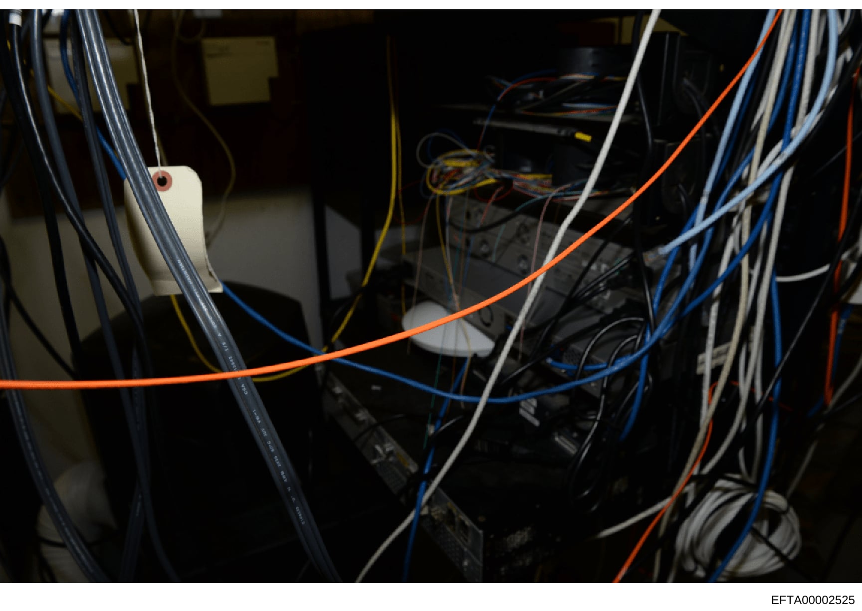 This is a photograph of the interior of technical equipment or a server/networking cabinet, showing multiple colored cables (orange, blue, white, yellow) connected to various black electronic devices and components. The image appears to be documentat