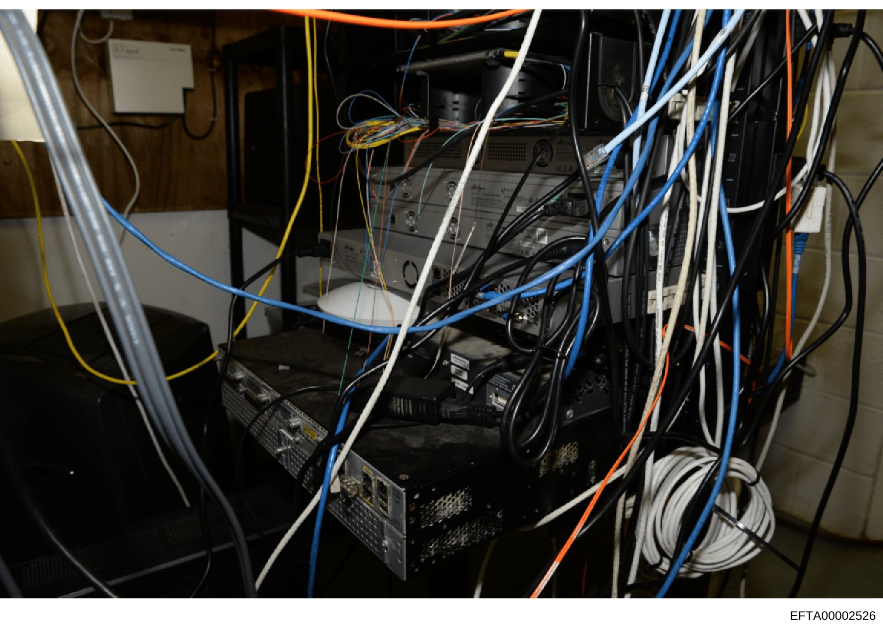 This is a photograph of a server room or network equipment closet showing multiple computer servers, networking hardware, and extensive cabling in various colors (blue, yellow, orange, white, black). The image documents the physical infrastructure an