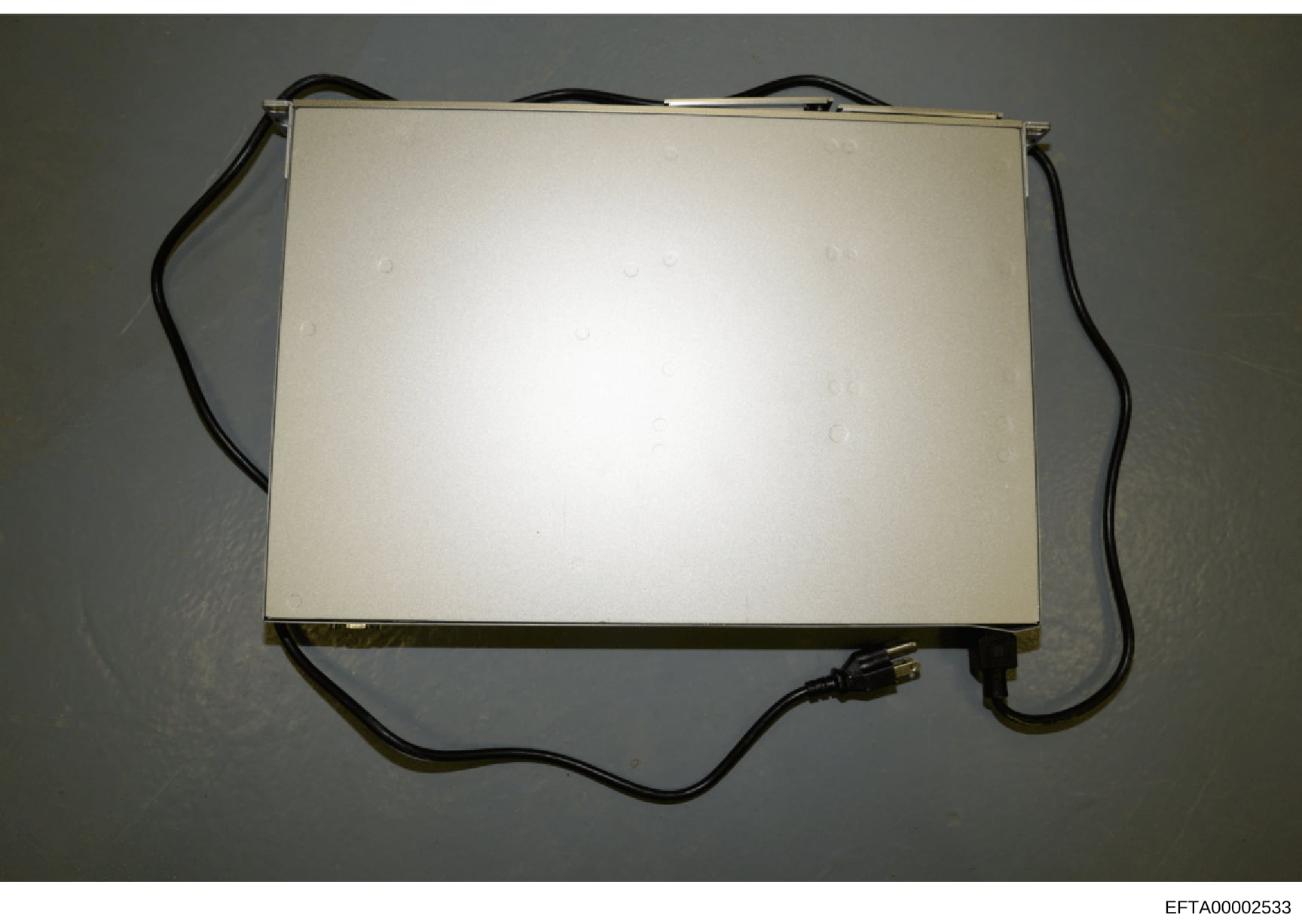 This is a photograph of a rectangular electronic device with a cream/beige colored top surface and black power cord attached. The device appears to be some form of lighting equipment or electronic apparatus, photographed against a gray wall. The imag