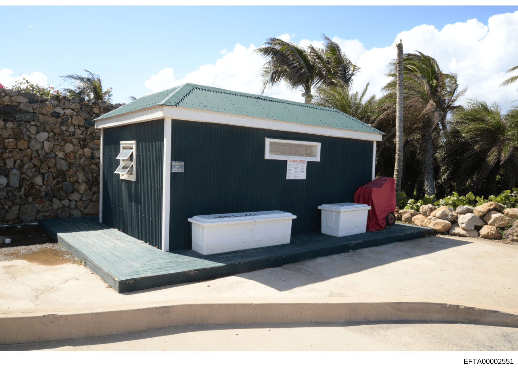 This photograph shows a dark-colored beach structure or facility with a turquoise roof, featuring two white storage containers on an elevated platform with wooden ramp access. The building is located in a tropical setting with palm trees and stone wa